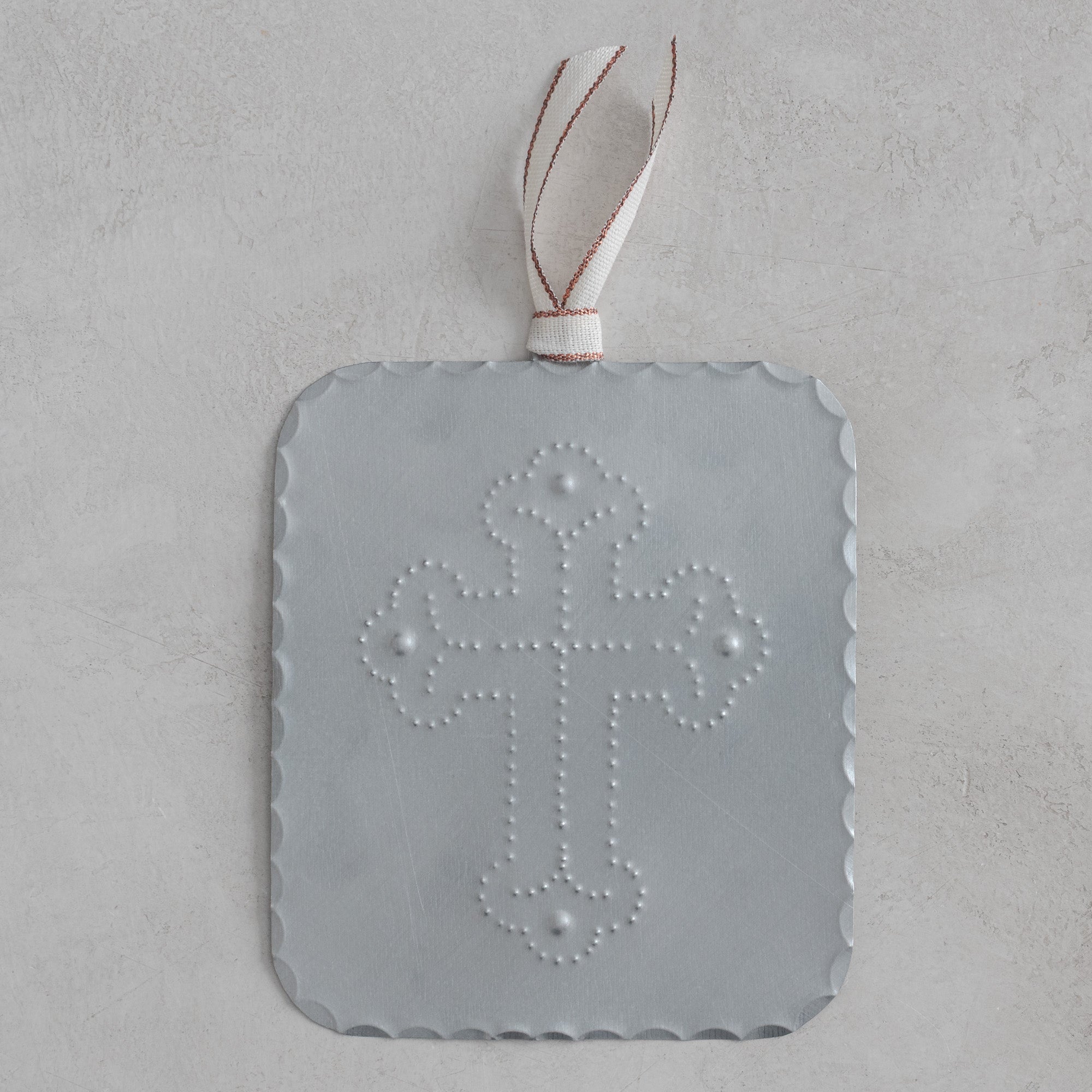 Hand-punched tin cross milagro with scalloped edges, finished in a soft matte silver tone, hanging from a cream ribbon with copper metallic edging on a light textured background.