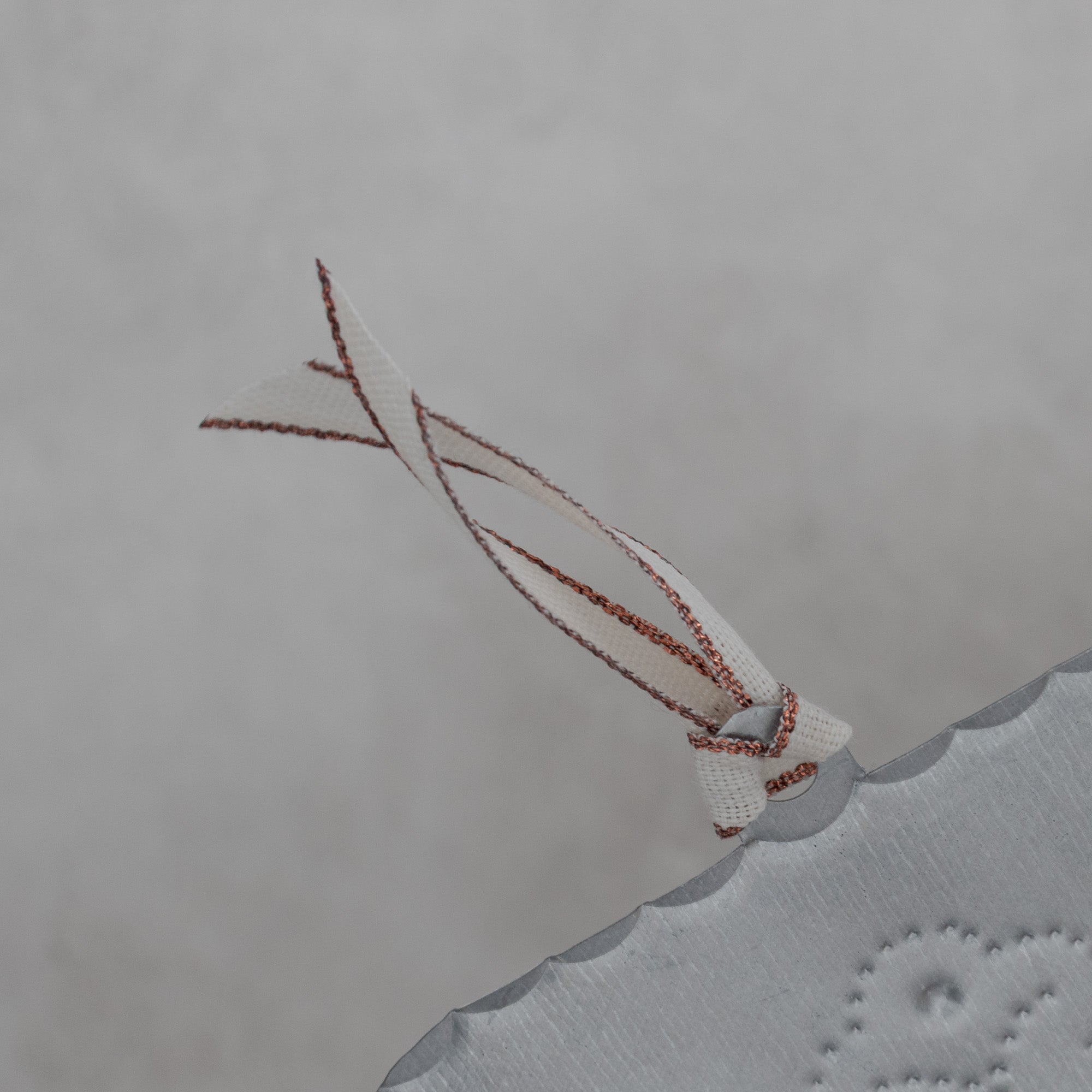 Back view detail of a hand-punched tin cross milagro showing the smooth tin surface, scalloped edge, and attached cream ribbon with copper metallic edging.