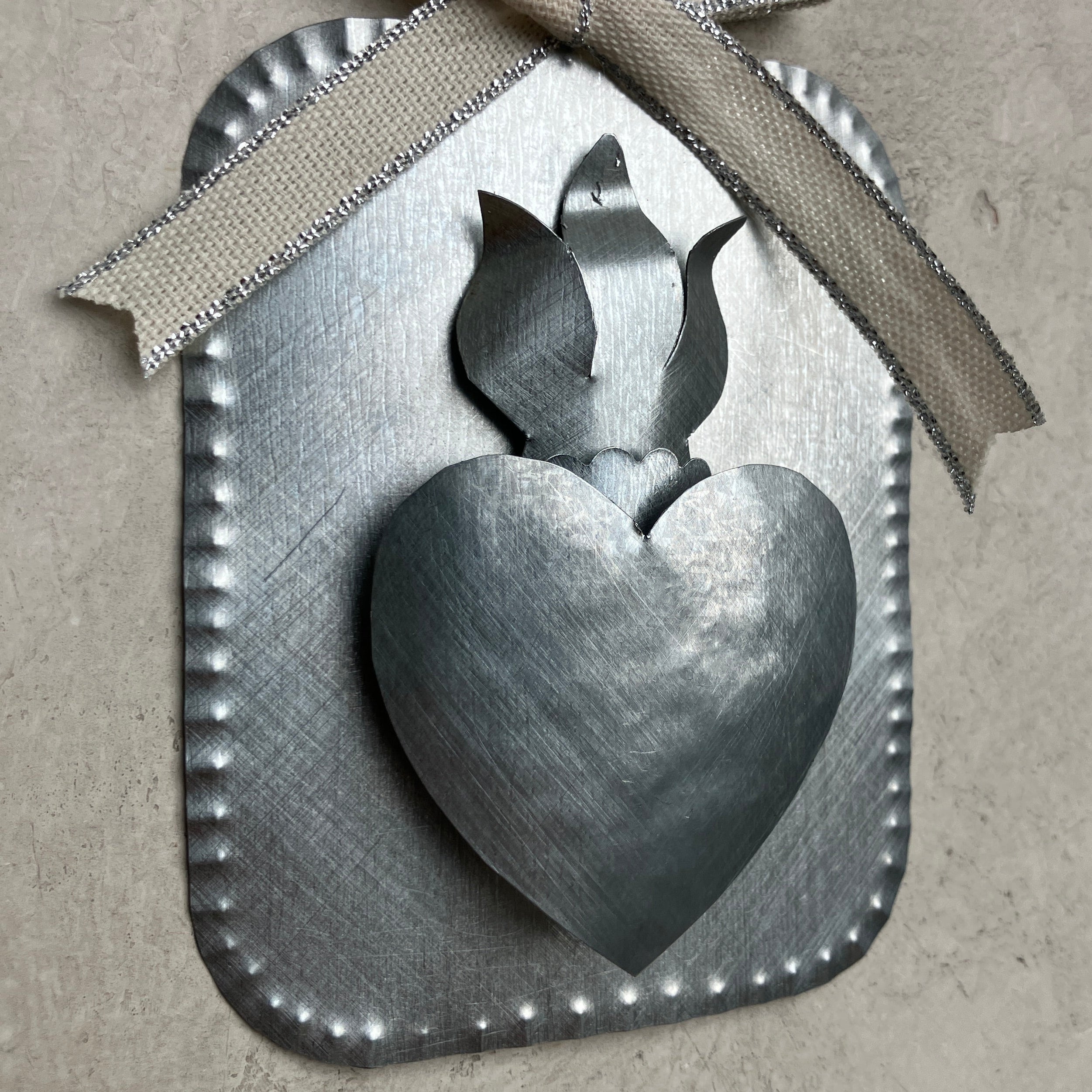 Close-up of a raised tin Sacred Heart ornament showing the dimensional heart and flame, scalloped edges, and a cream ribbon with metallic trim against a textured gray surface.