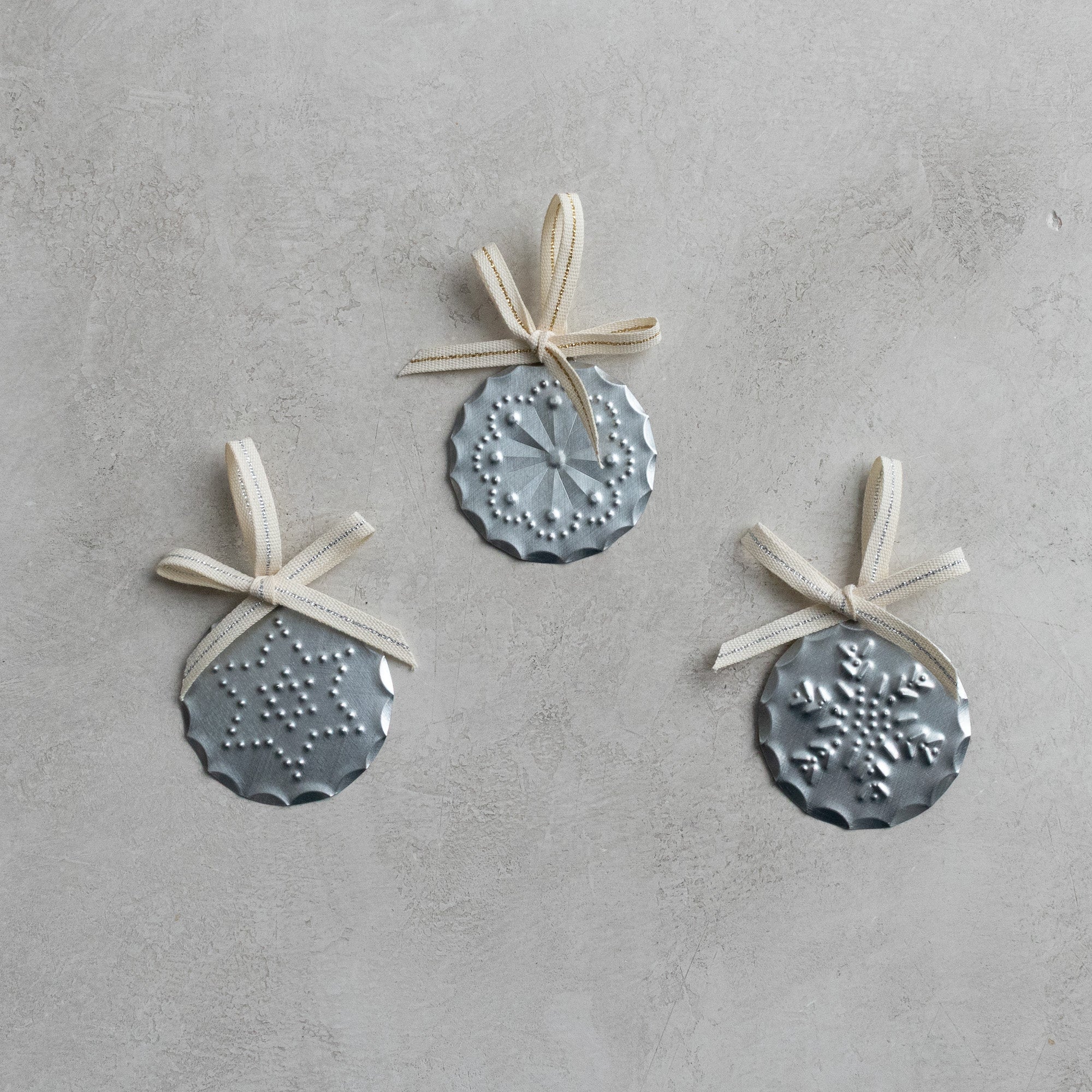 Three hand-punched tin ornaments with ribbon bows arranged on a neutral stone background.