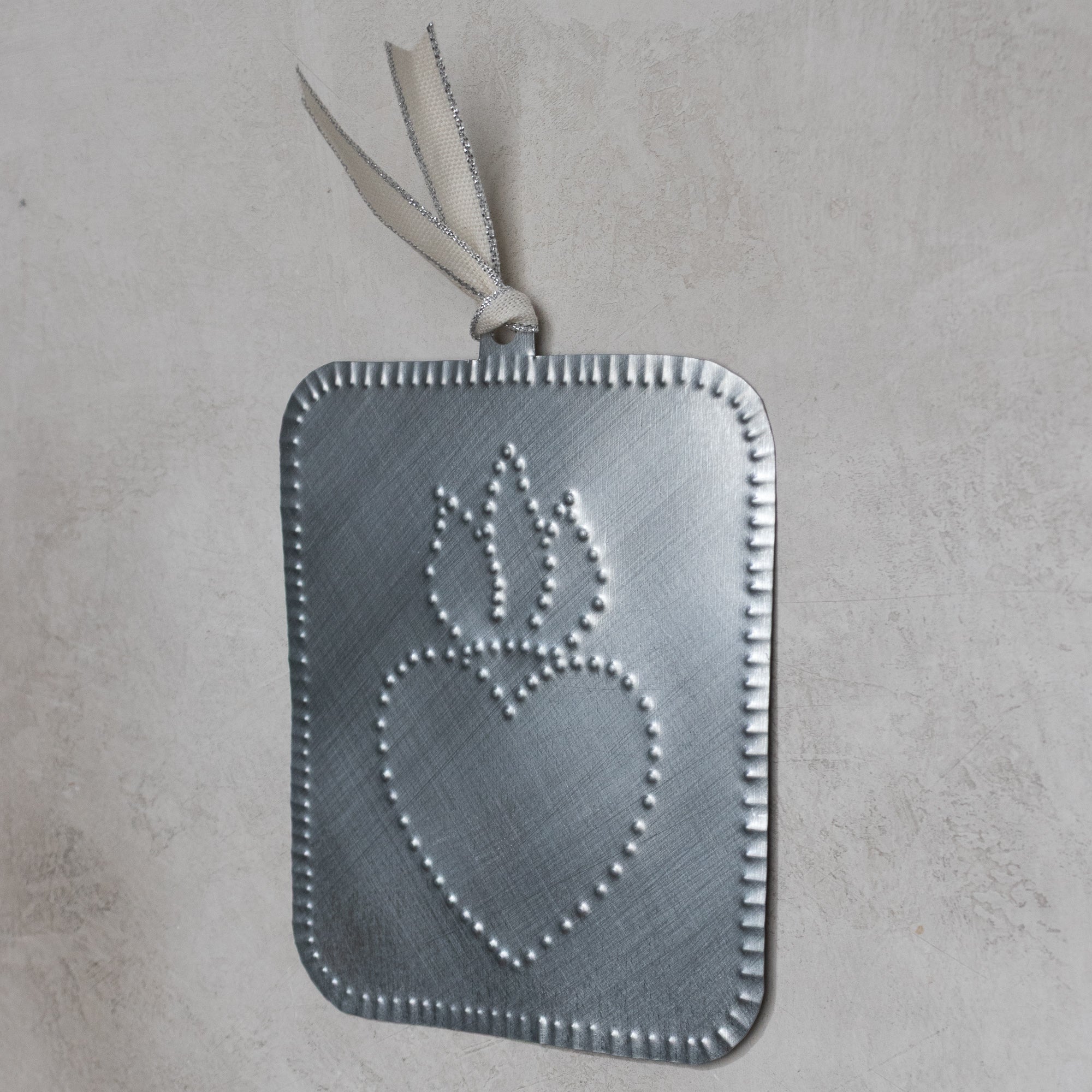 Handmade tin ornament with a punched flaming heart design, shown at an angle against a light stone-textured background.