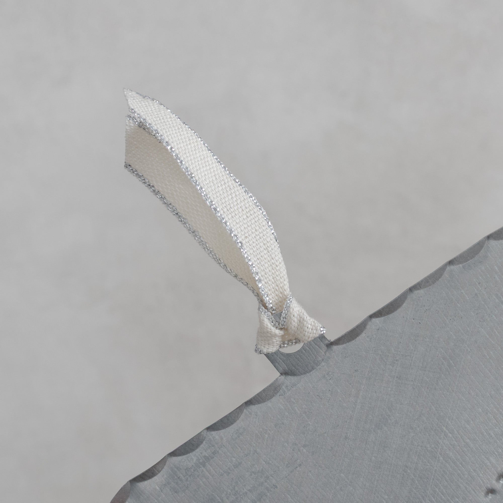 Back view detail of a hand-punched tin ojo milagro showing the smooth tin surface, scalloped edge, and cream ribbon with silver metallic edging.