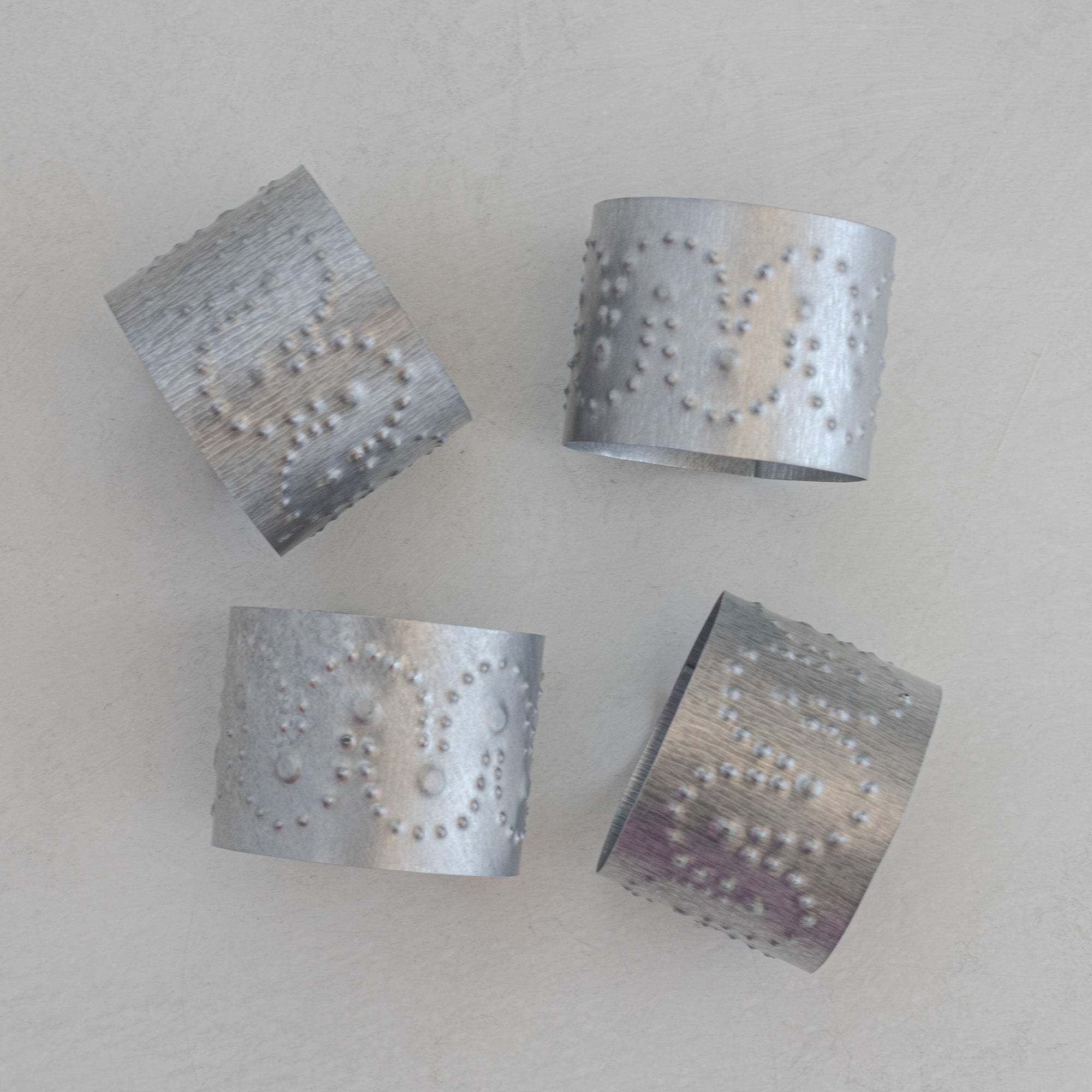 Set of four Arco hand-punched tin napkin rings scattered on a light plaster surface, showing arco motif with dot accents in natural tin.