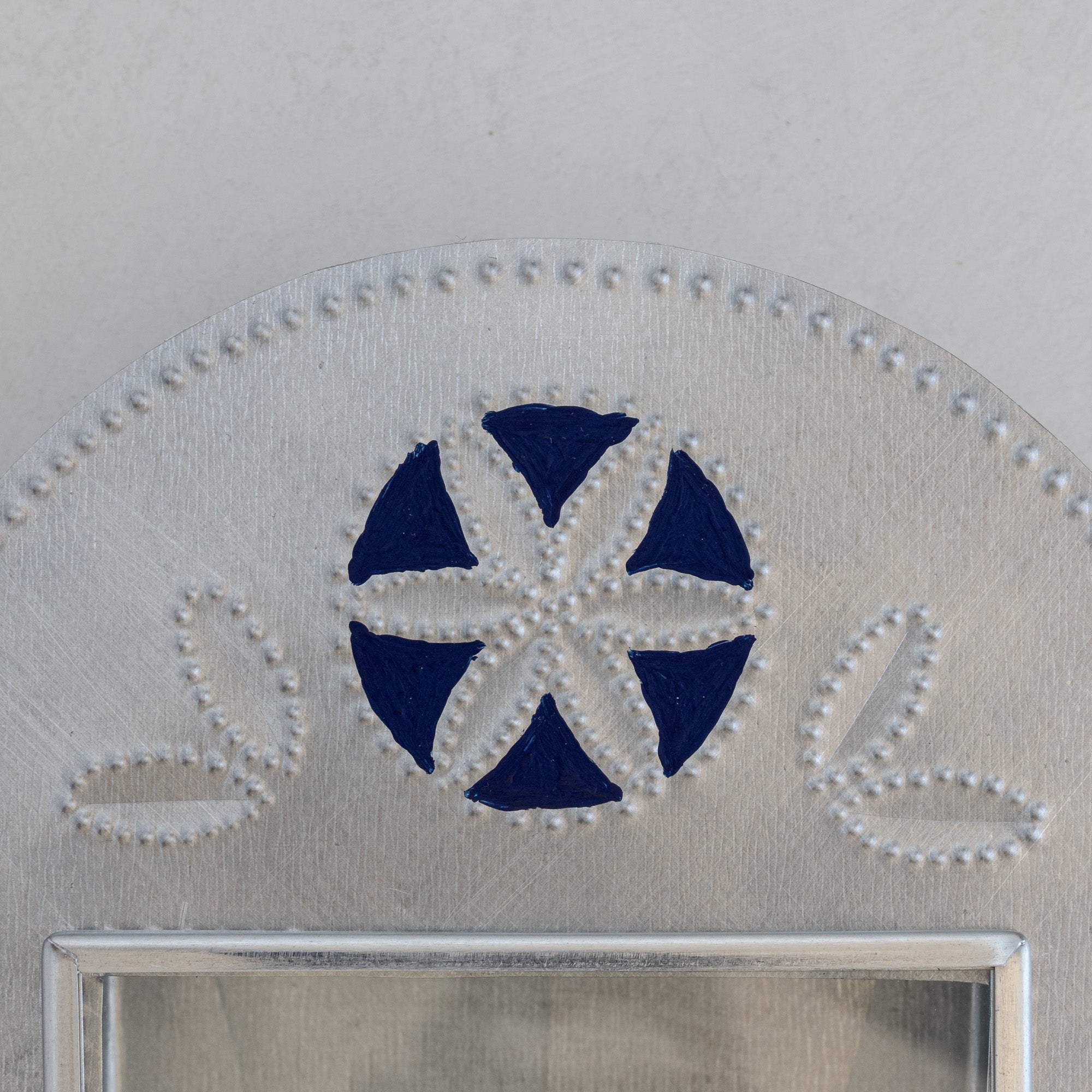Close-up of Nicho de Recuerdo crest showing hand-punched floral motif painted in Prussian blue on natural tin, with dot-punched border and leaf forms flanking.