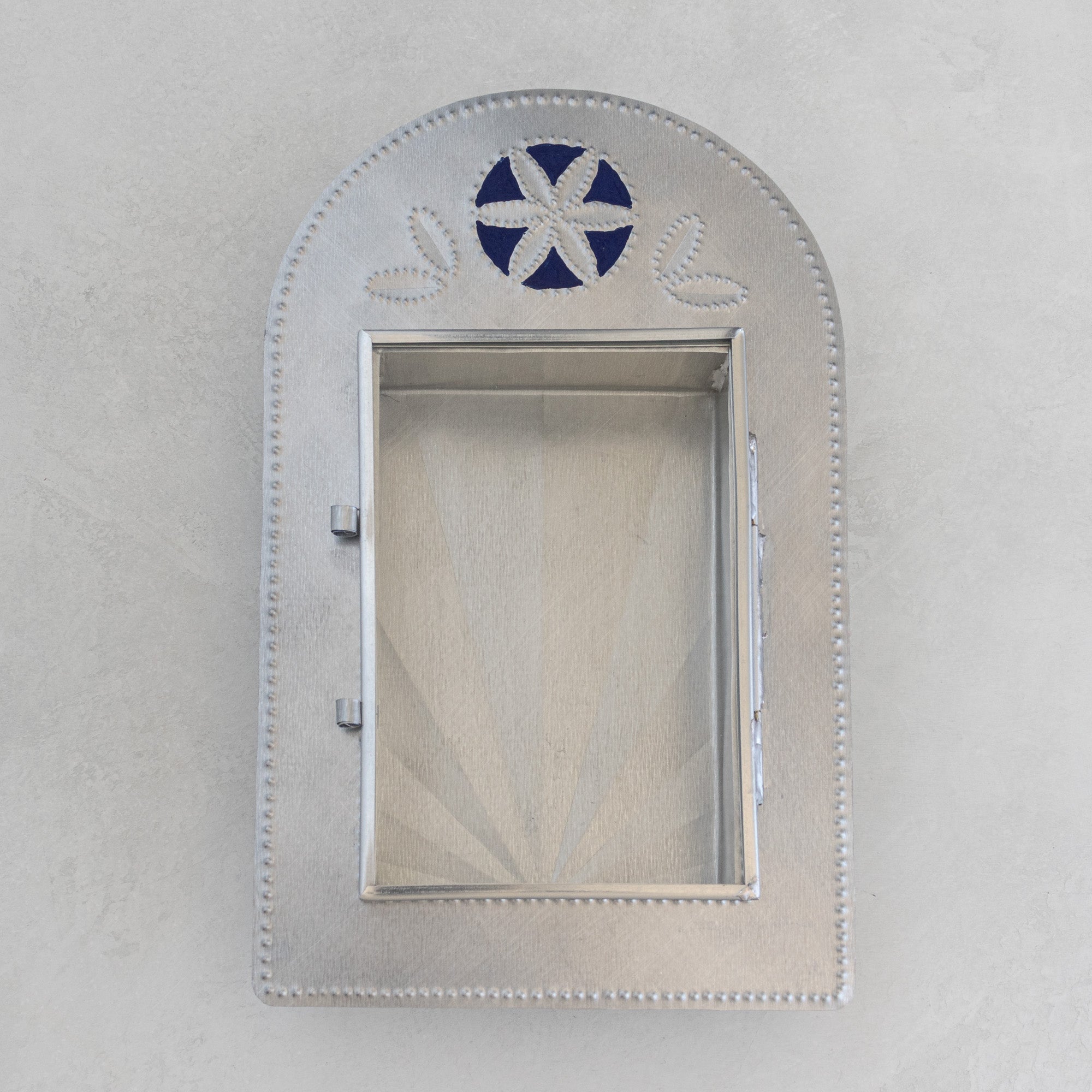 Nicho de Recuerdo laid flat, door closed, showing full arched tin body, Prussian blue painted floral crest motif, dot-punched border, and handle.