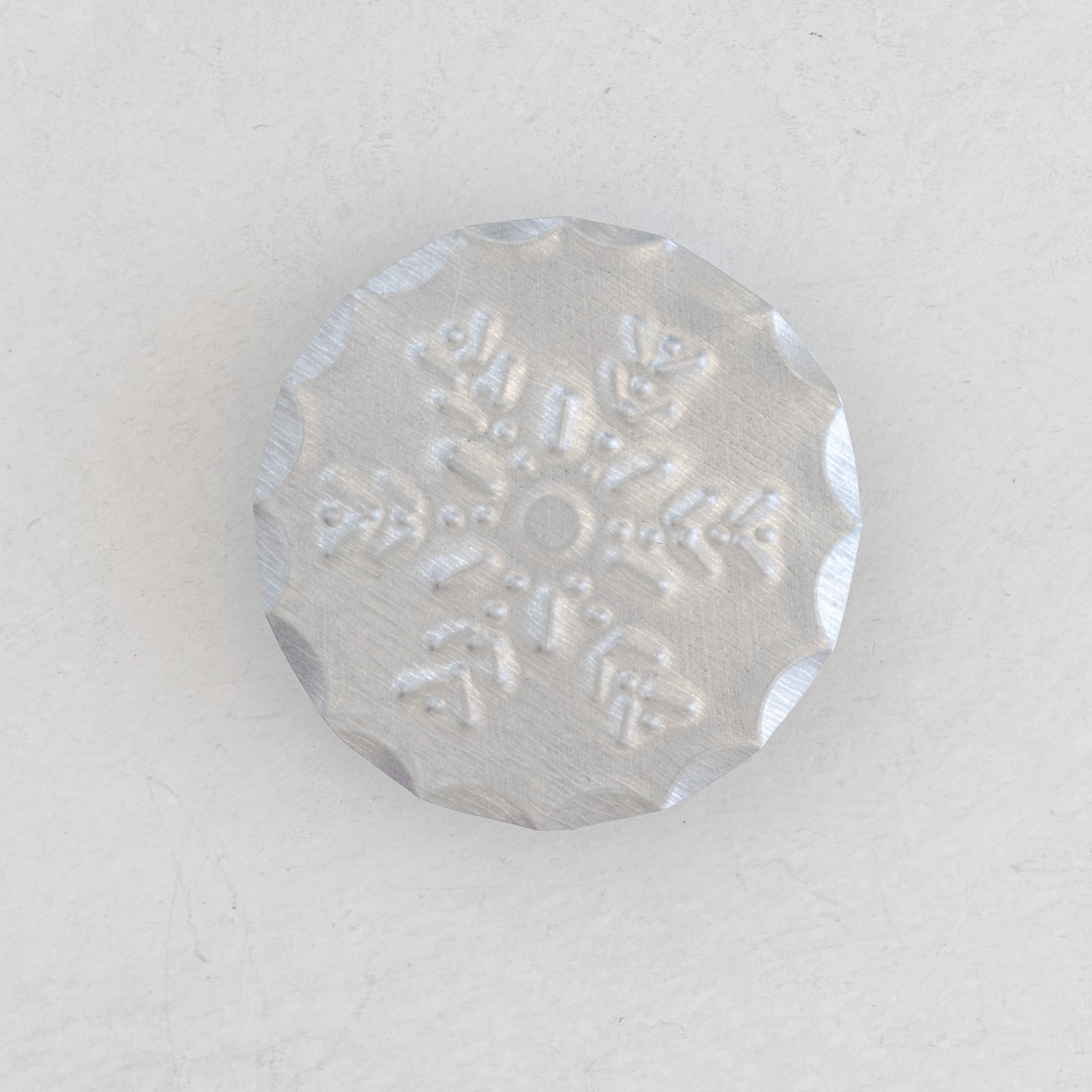 Nieve Bandana Slide front view showing hand-punched snowflake motif on a crimped-edge circular tin disc, natural tin, light plaster surface.