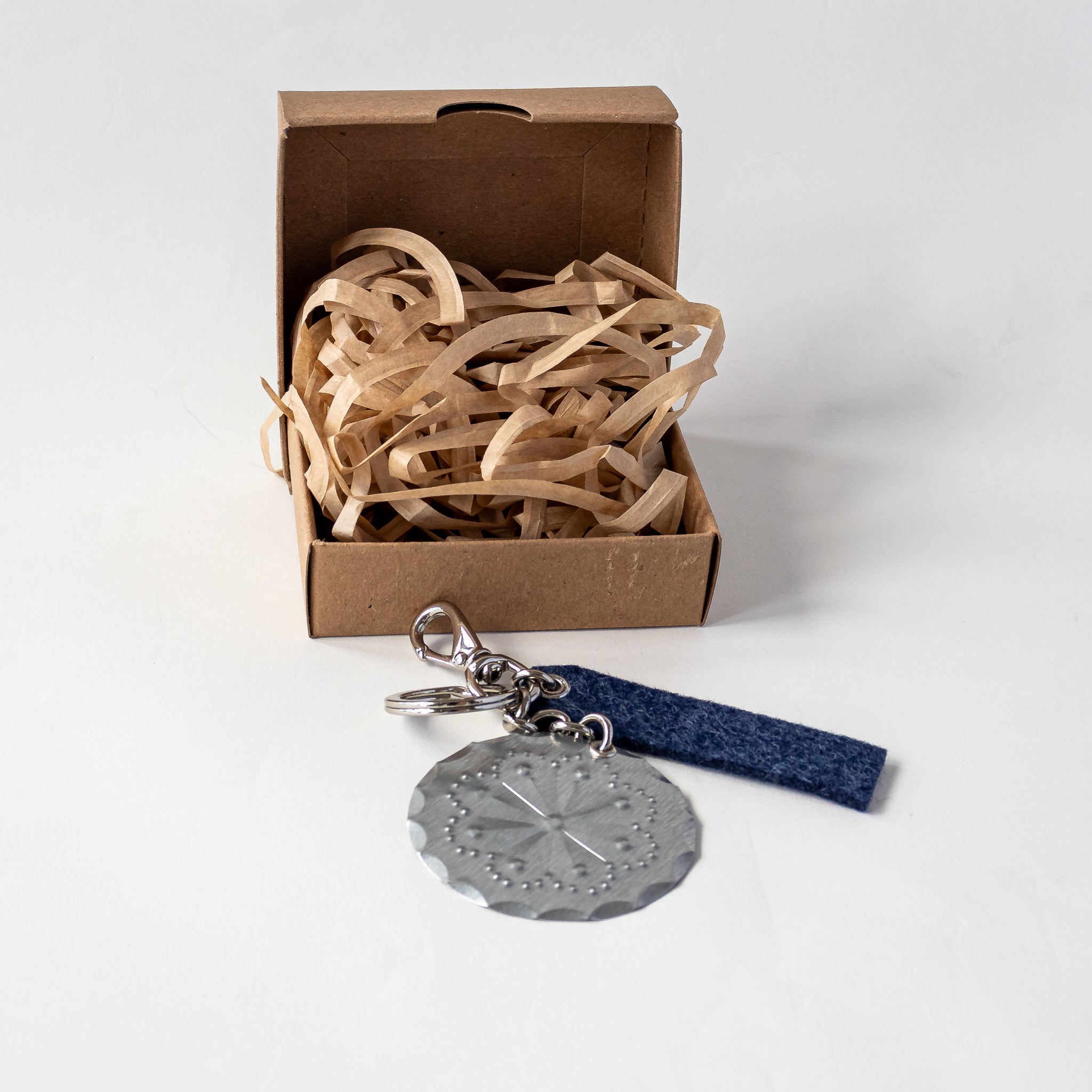 Accent Lever Bag Charm with indigo felt tag, shown with kraft jewelry gift box and paper shred.
