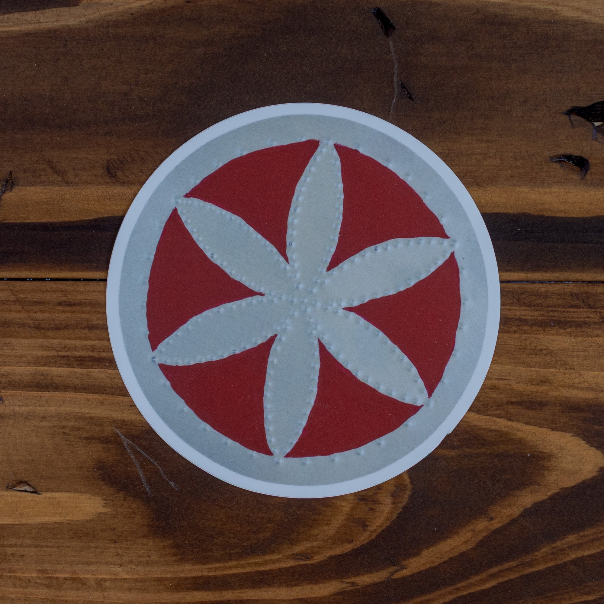 Flor sticker close up on dark wood grain, showing punched tin petal design in silver on deep red circle