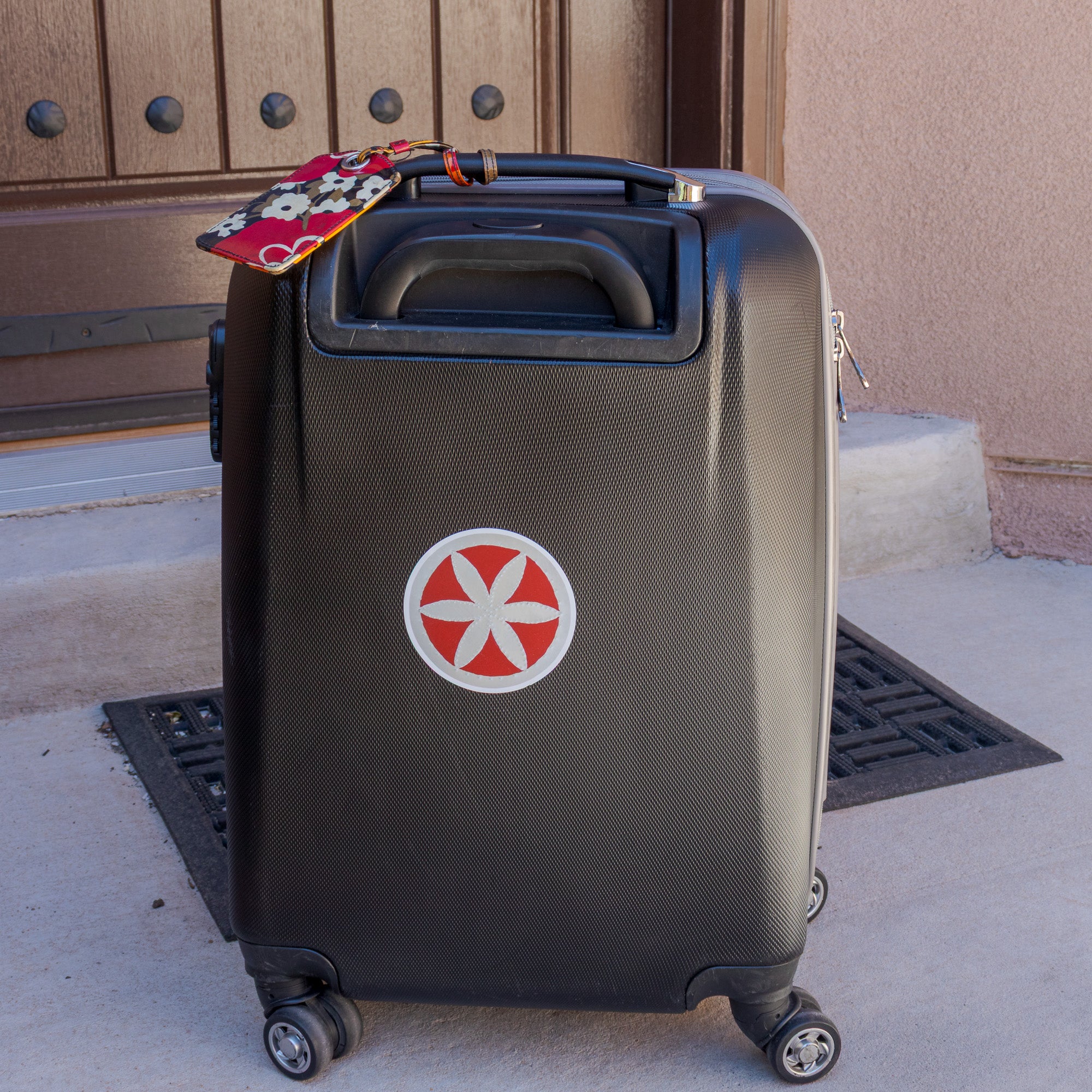 Flor sticker on a dark rolling suitcase outside an adobe building, luggage tag visible at the handle