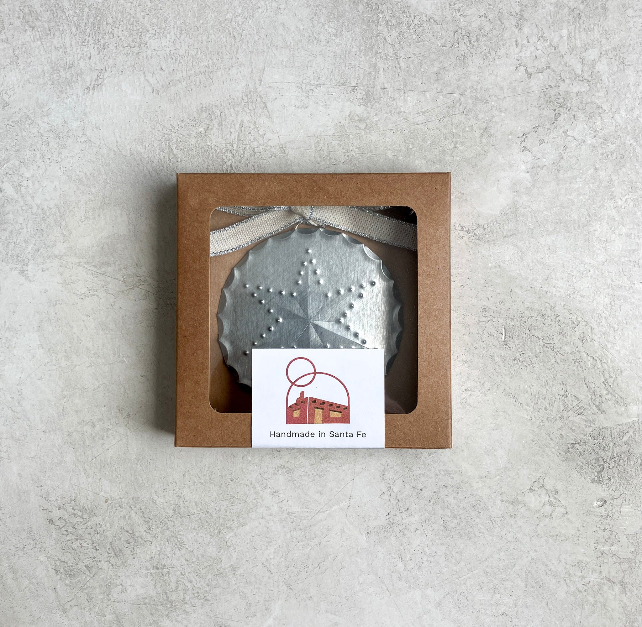 The Luz Estrella tin ornament with a six-pointed punched star design, packaged in a kraft window box with a metallic-edged ribbon .