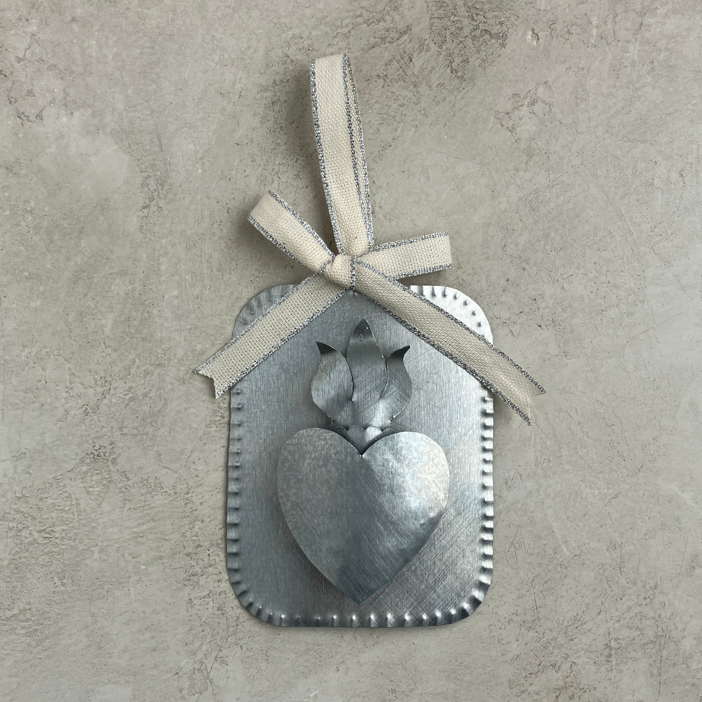 A raised tin Sacred Heart ornament with a dimensional heart and flame sits centered on a textured neutral background. It is finished with a cream Studio Carta ribbon tied in a bow at the top.
