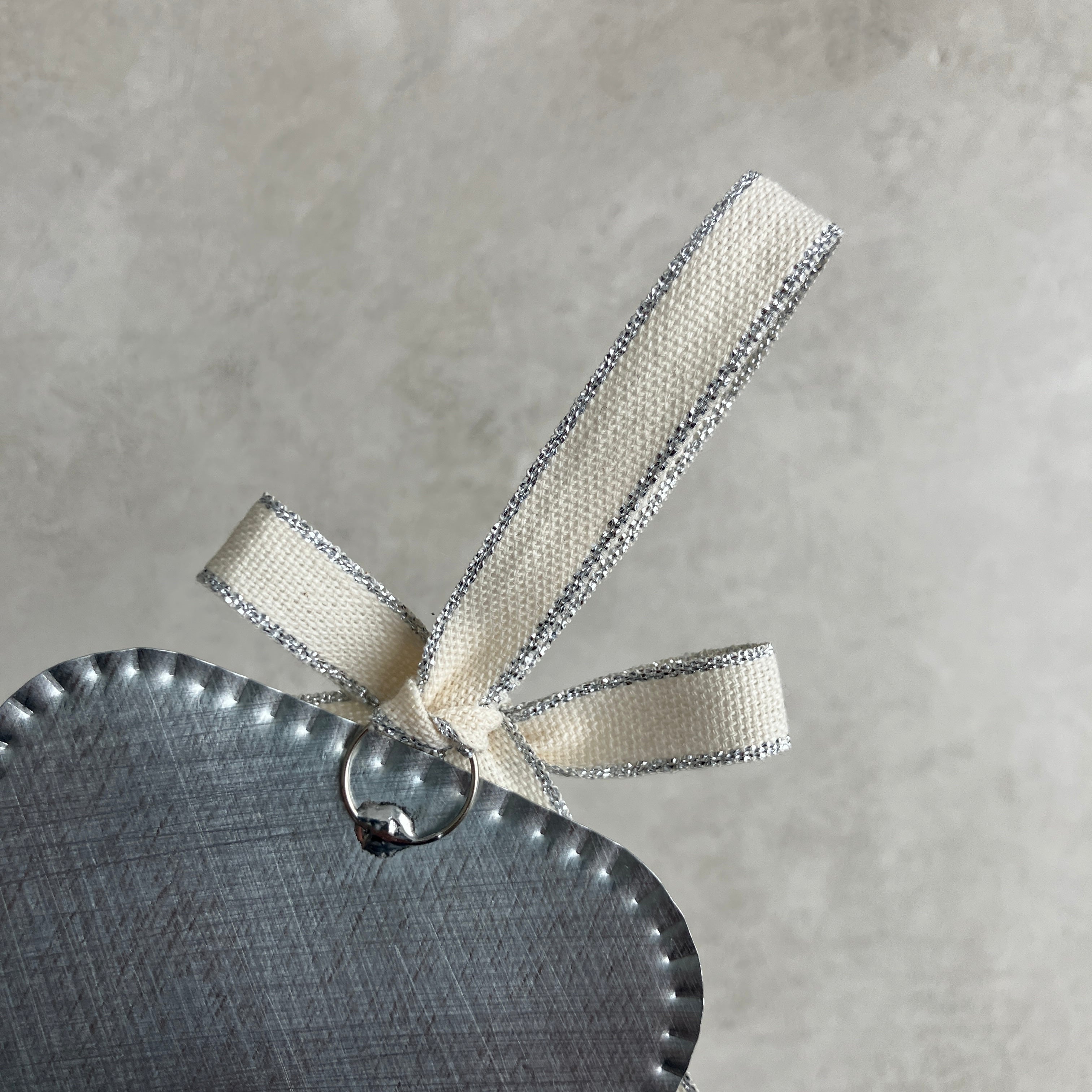 Close-up of the top of a raised tin Sacred Heart ornament, showing the scalloped edge, hanging ring, and a cream ribbon with metallic trim against a soft gray background.