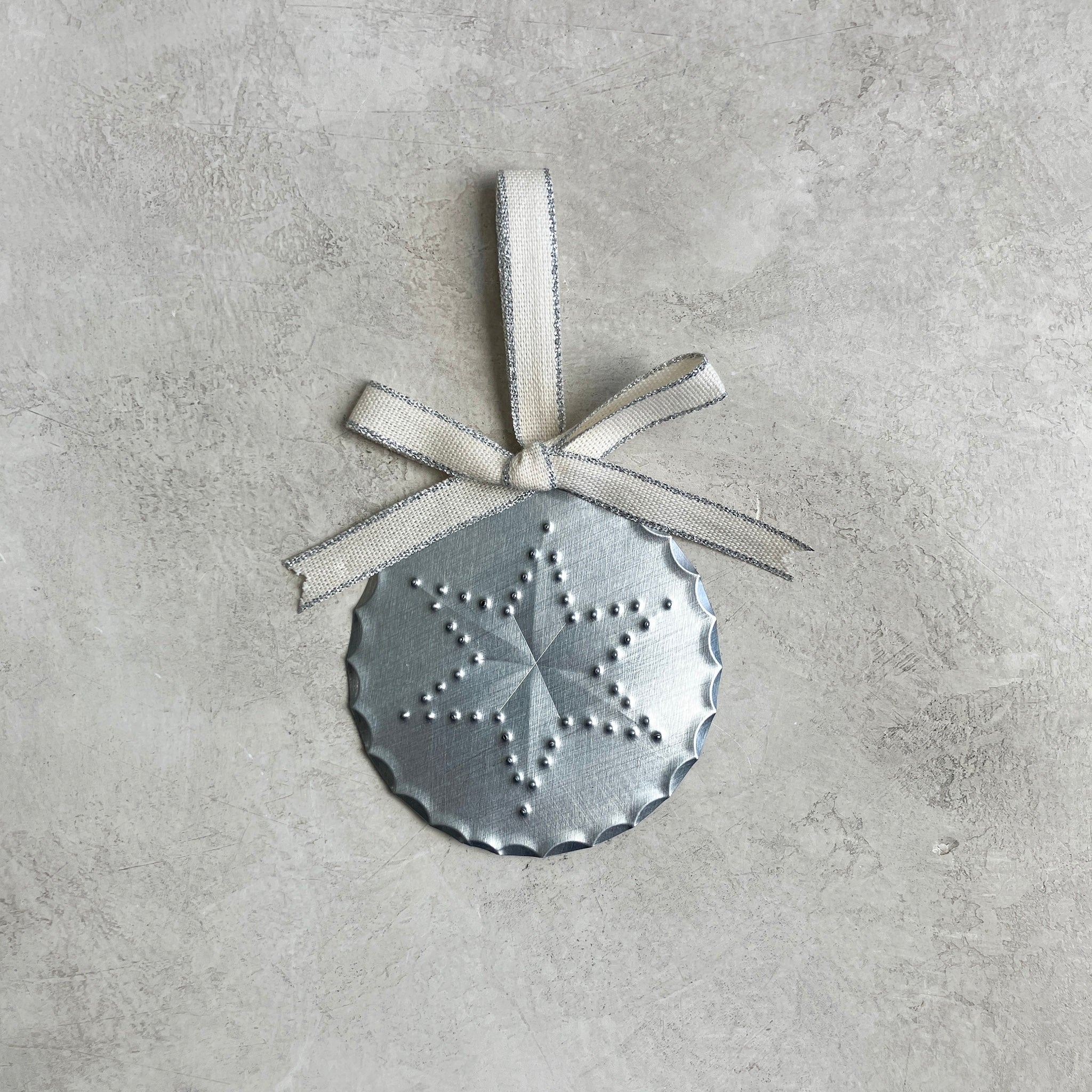 A round tin ornament with a hand-punched six-pointed star design, scalloped edges, and a metallic-edged cream ribbon bow, photographed on a light stone background.