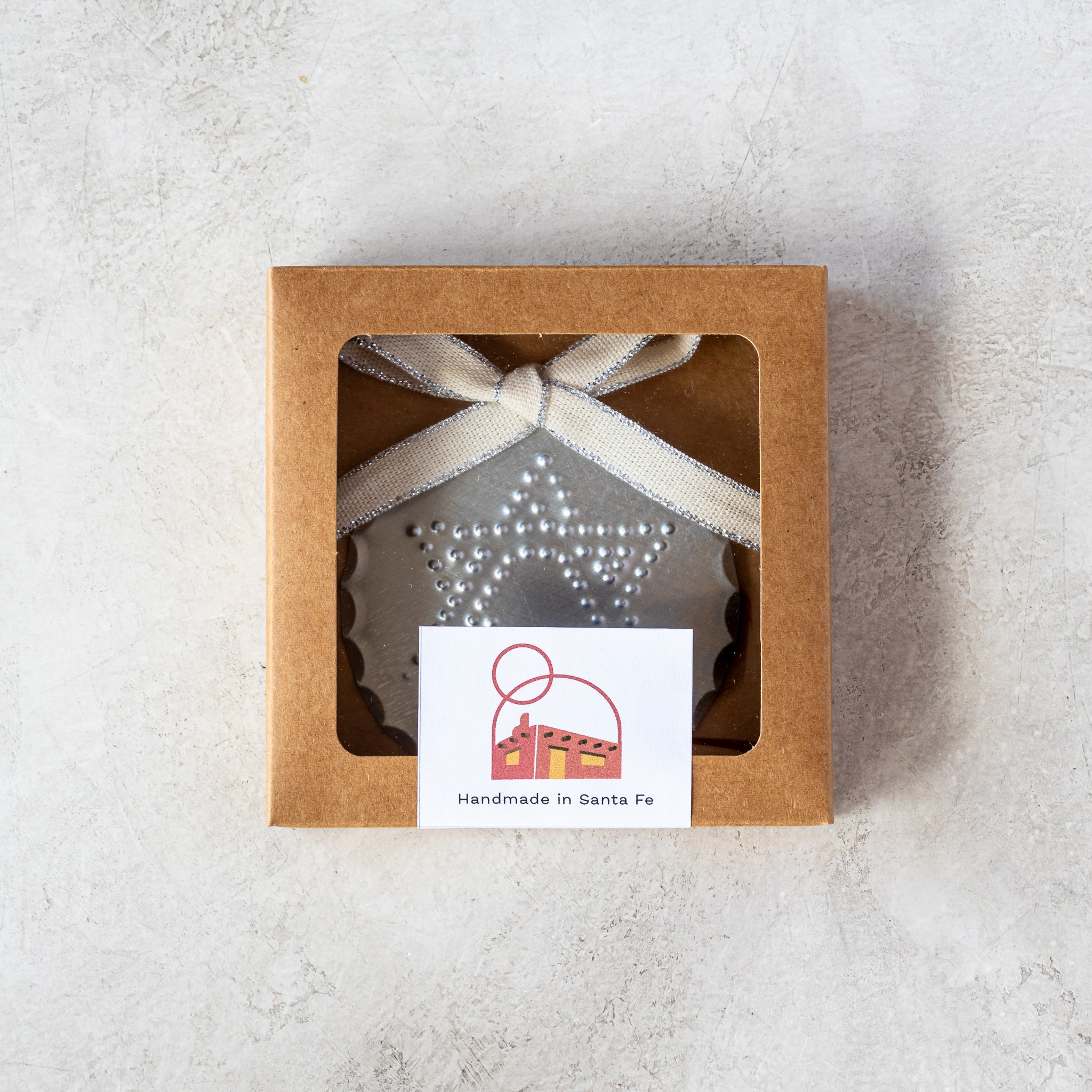 Gift-ready 3” tin Luz Estrella Ornament in kraft window box with Studio Carta ribbon.