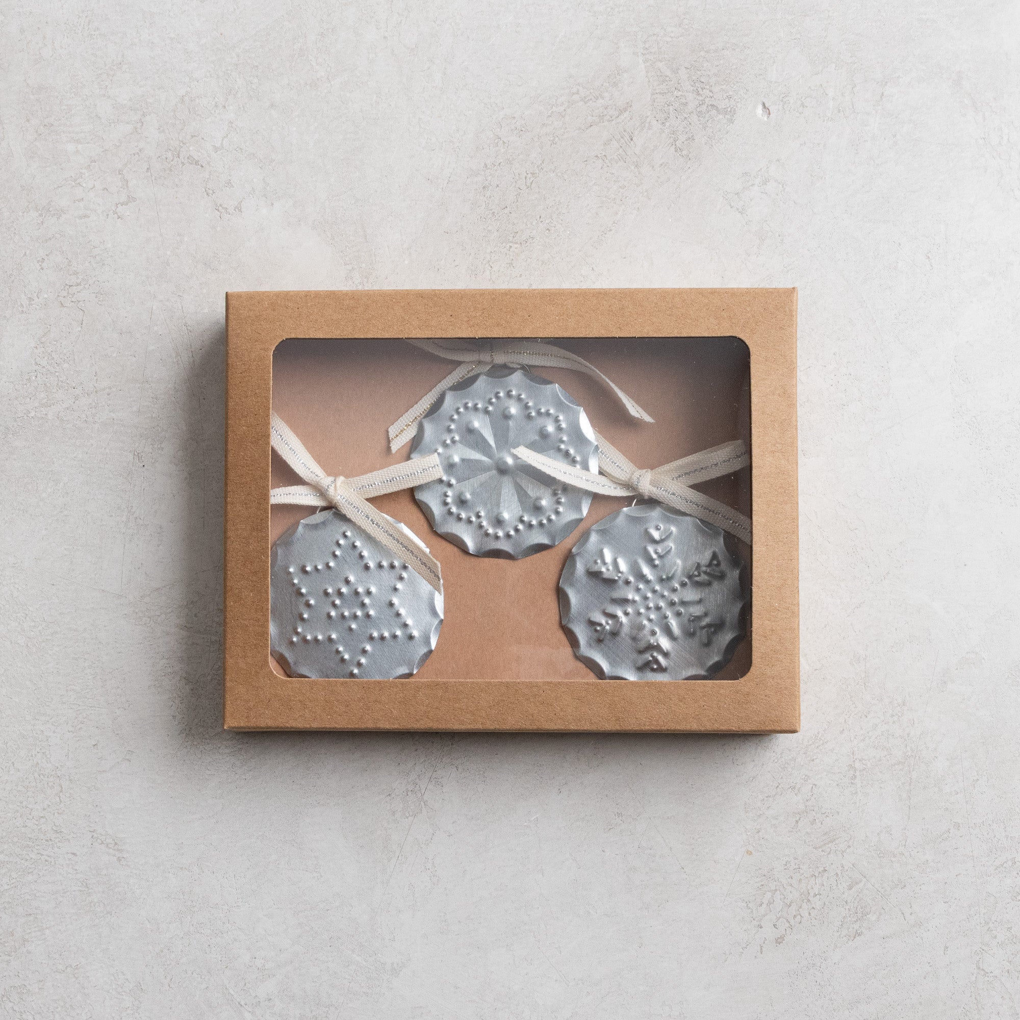 Boxed set of three hand-punched tin ornaments with ribbon bows, shown in a kraft window box on a neutral stone background.