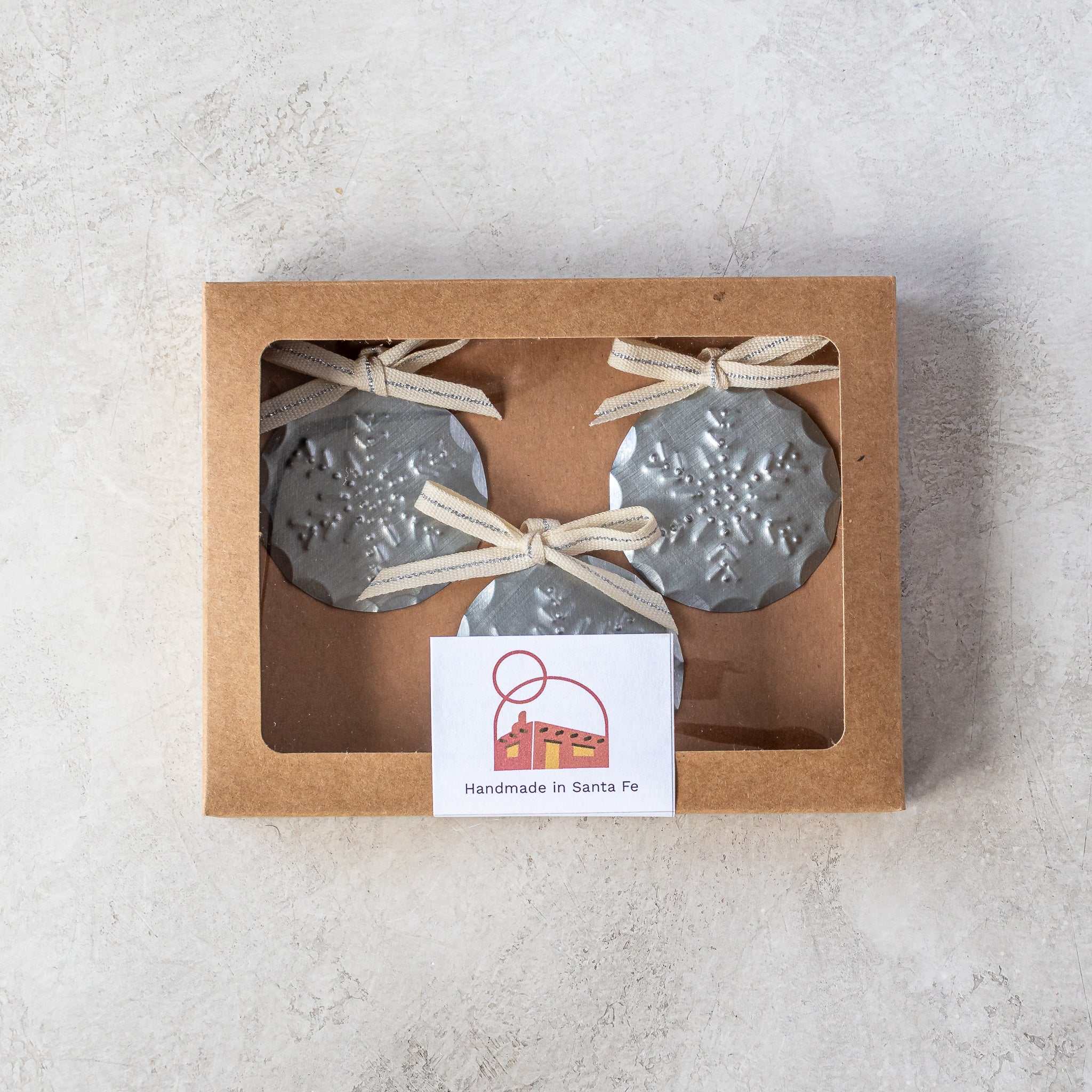 Gift-ready set of three 2” tin Luz Nieve Mini Ornaments in kraft window box with Studio Carta ribbon.
