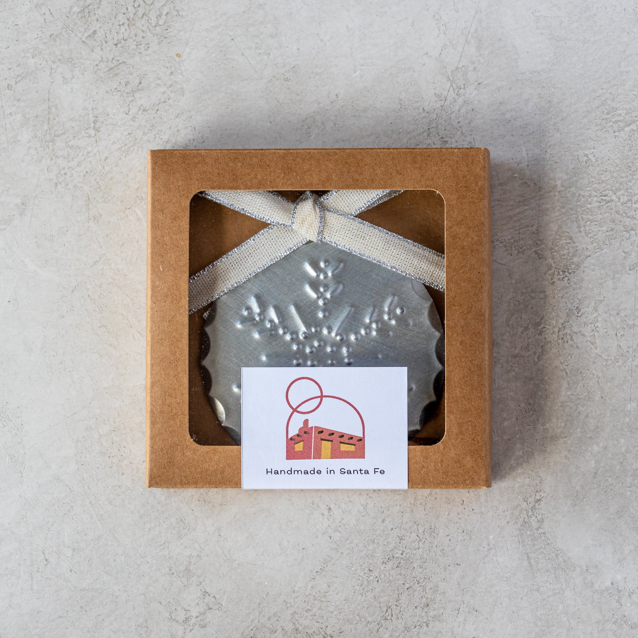 Gift-ready 3” tin Luz Nieve Ornament in kraft window box with Studio Carta ribbon.