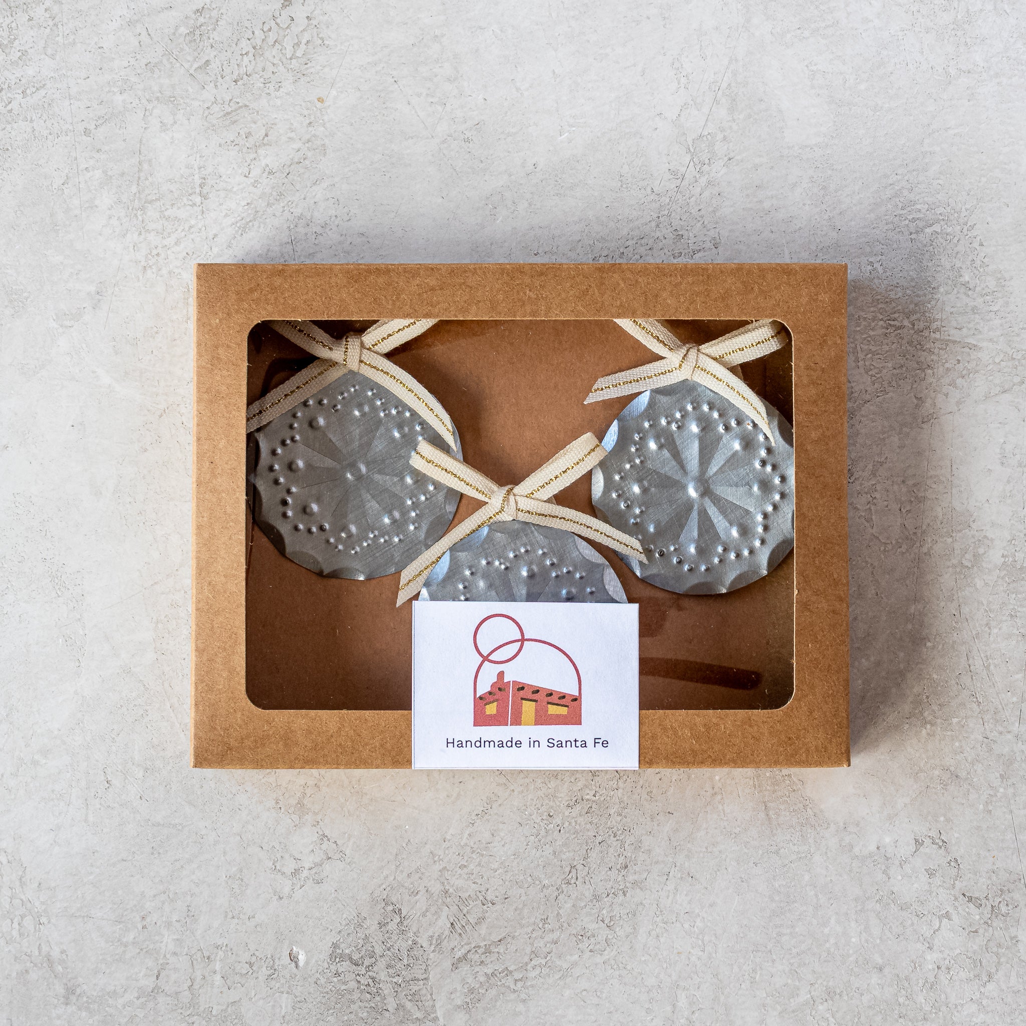Gift-ready set of three 2” tin Luz Roseta Mini Ornaments in kraft window box with Studio Carta ribbon.