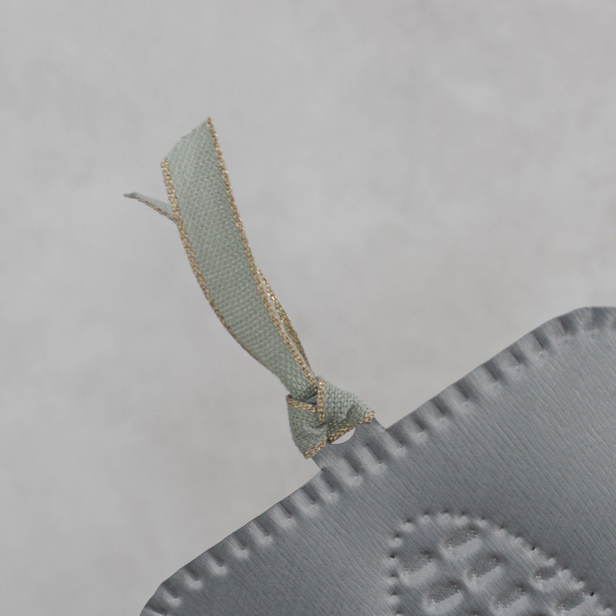 Back view detail of a hand-punched tin maize milagro showing the smooth tin surface, scalloped edge, and pale green ribbon with gold metallic edging.