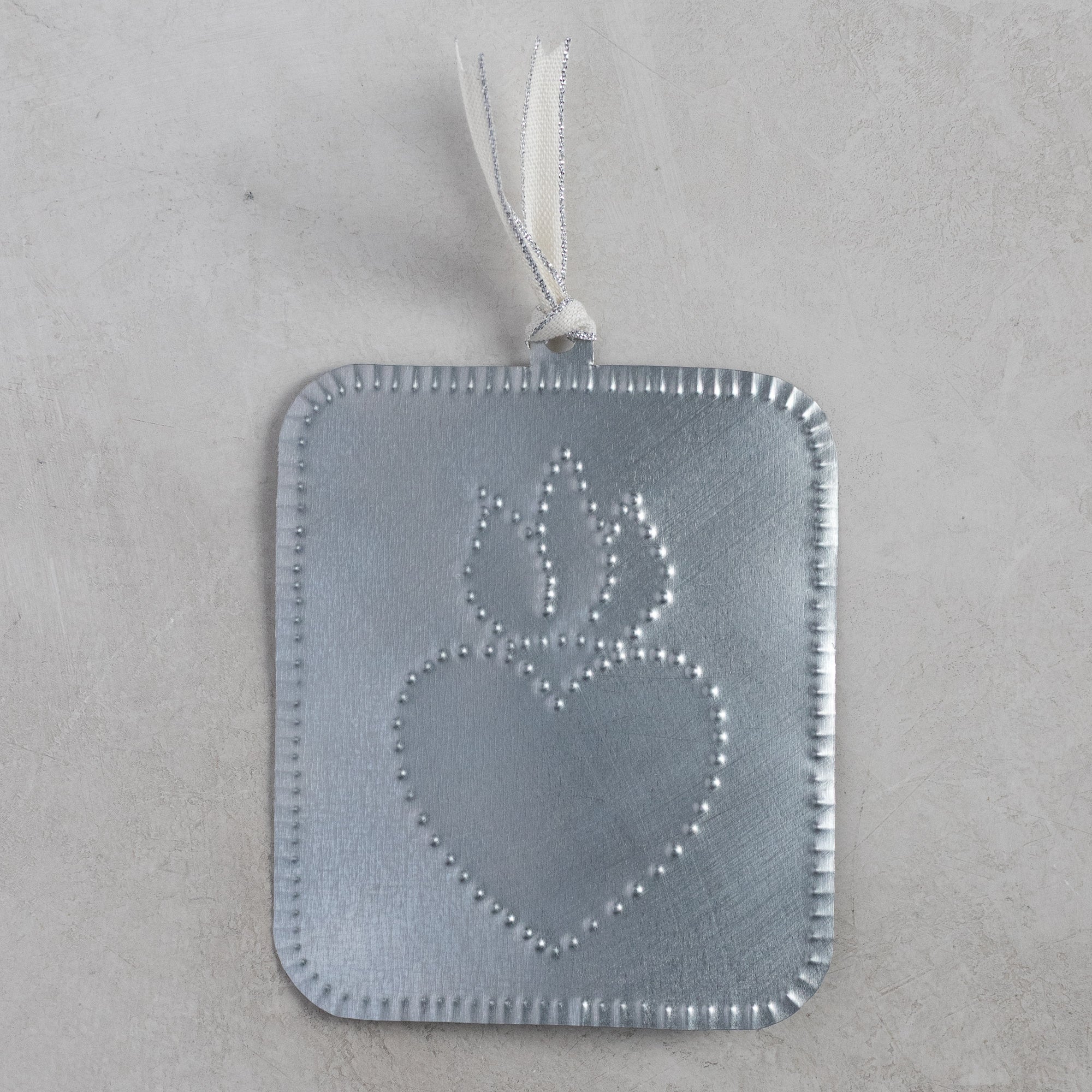 Handmade tin ornament with a punched flaming heart design, photographed flat against a light stone-textured background.