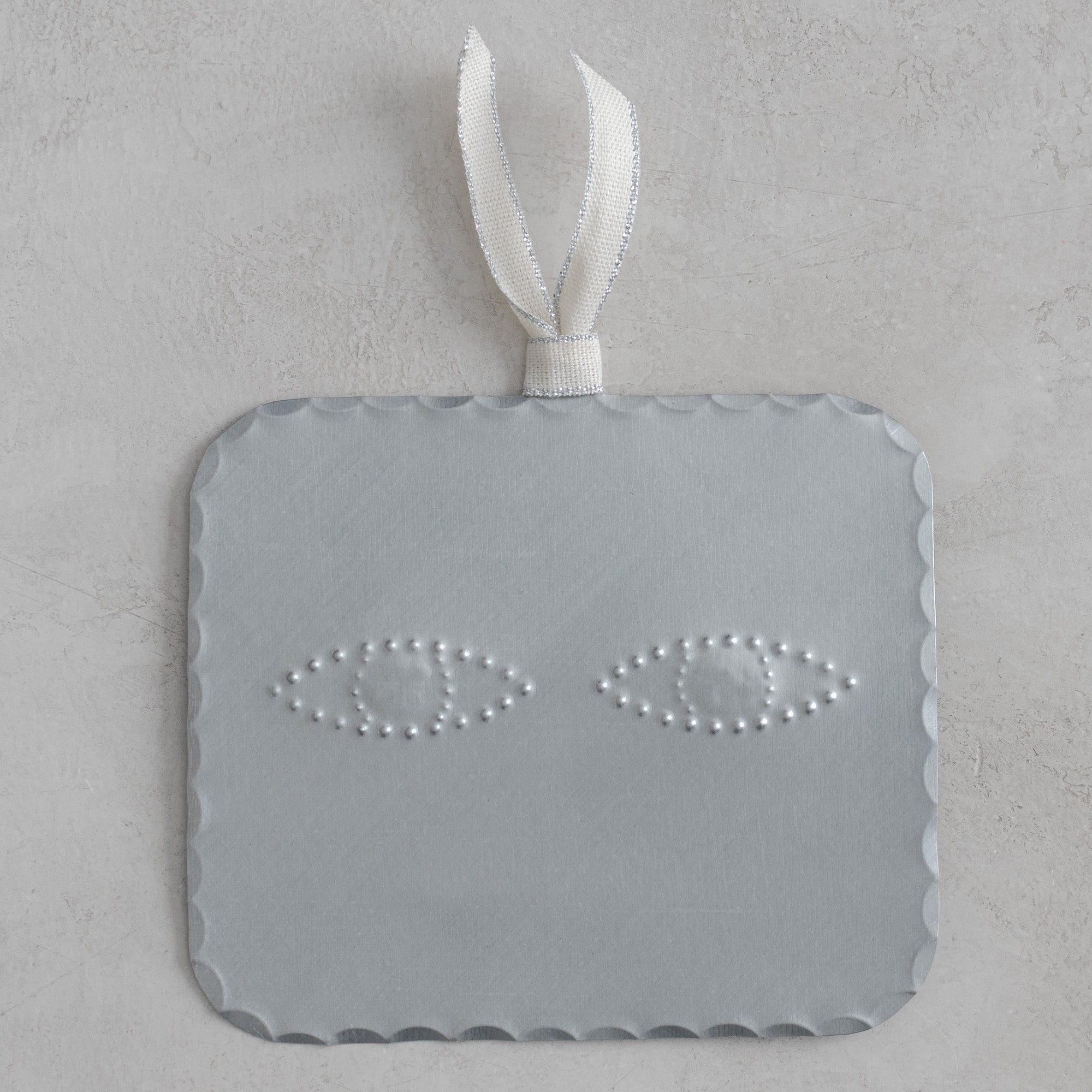 Hand-punched tin ojo (eye) milagro with scalloped edges, finished in a soft matte silver tone, hanging from a cream ribbon with silver metallic edging on a light textured background.
