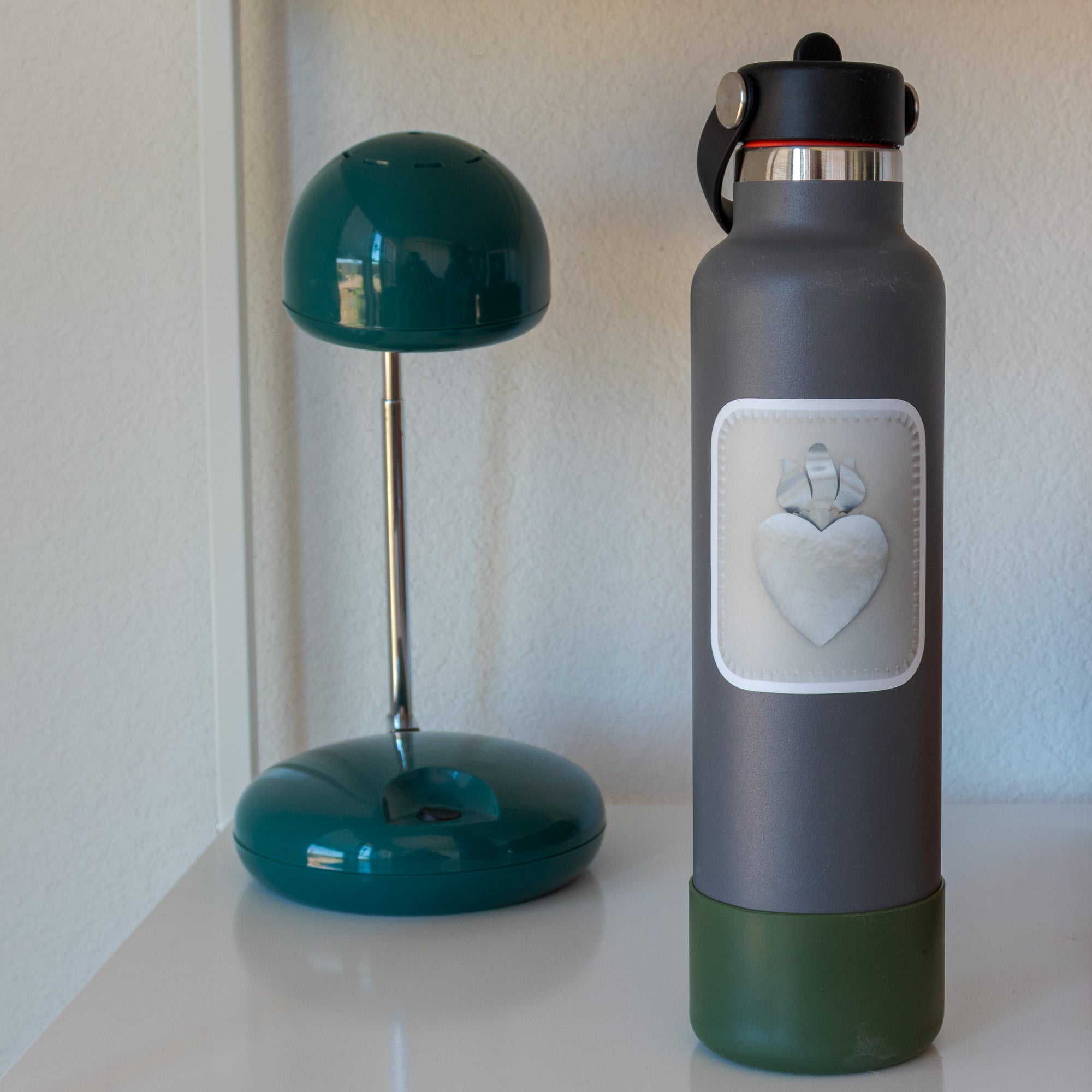 Sacred Heart sticker on a grey water bottle beside a teal dome lamp on a white shelf