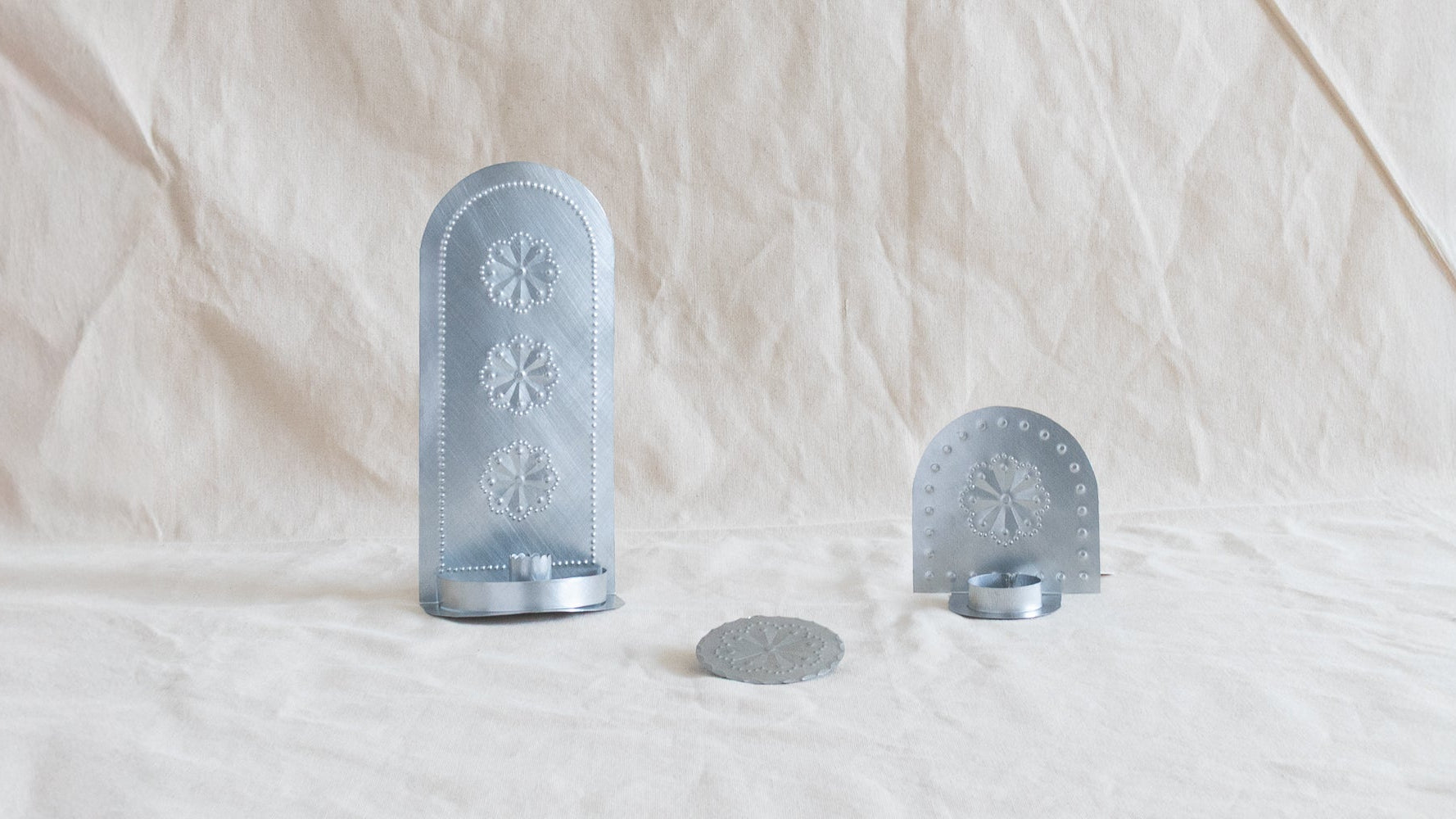 Three hand-punched tin pieces by Homekeep Studio arranged on a linen background — a tall Roseta taper candleholder, a smaller tealight candleholder, and a flat ornament disc, made in Santa Fe, New Mexico.