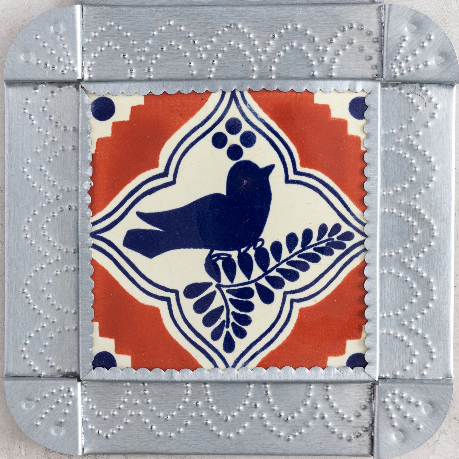 Closeup of a decorative tinwwork framed tile with a blue bird design on a terracotta and cream background, surrounded by gray border.
