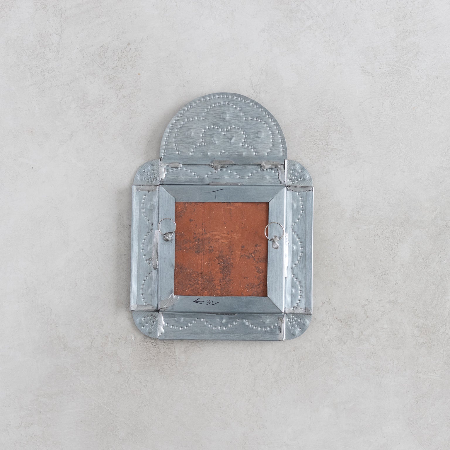 Back of a decorative tinwork framed tile wall decor with hangers on a light gray background.