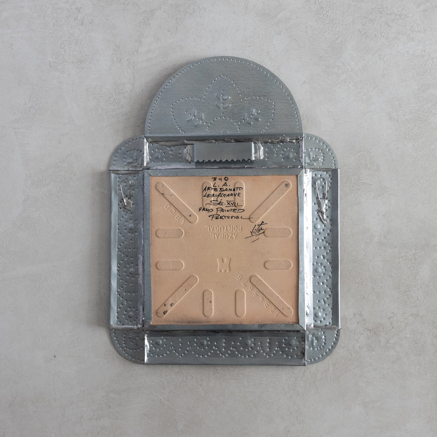 Back of a decorative  tinwork framed tile with sawtooth hangers and loops on a gray background