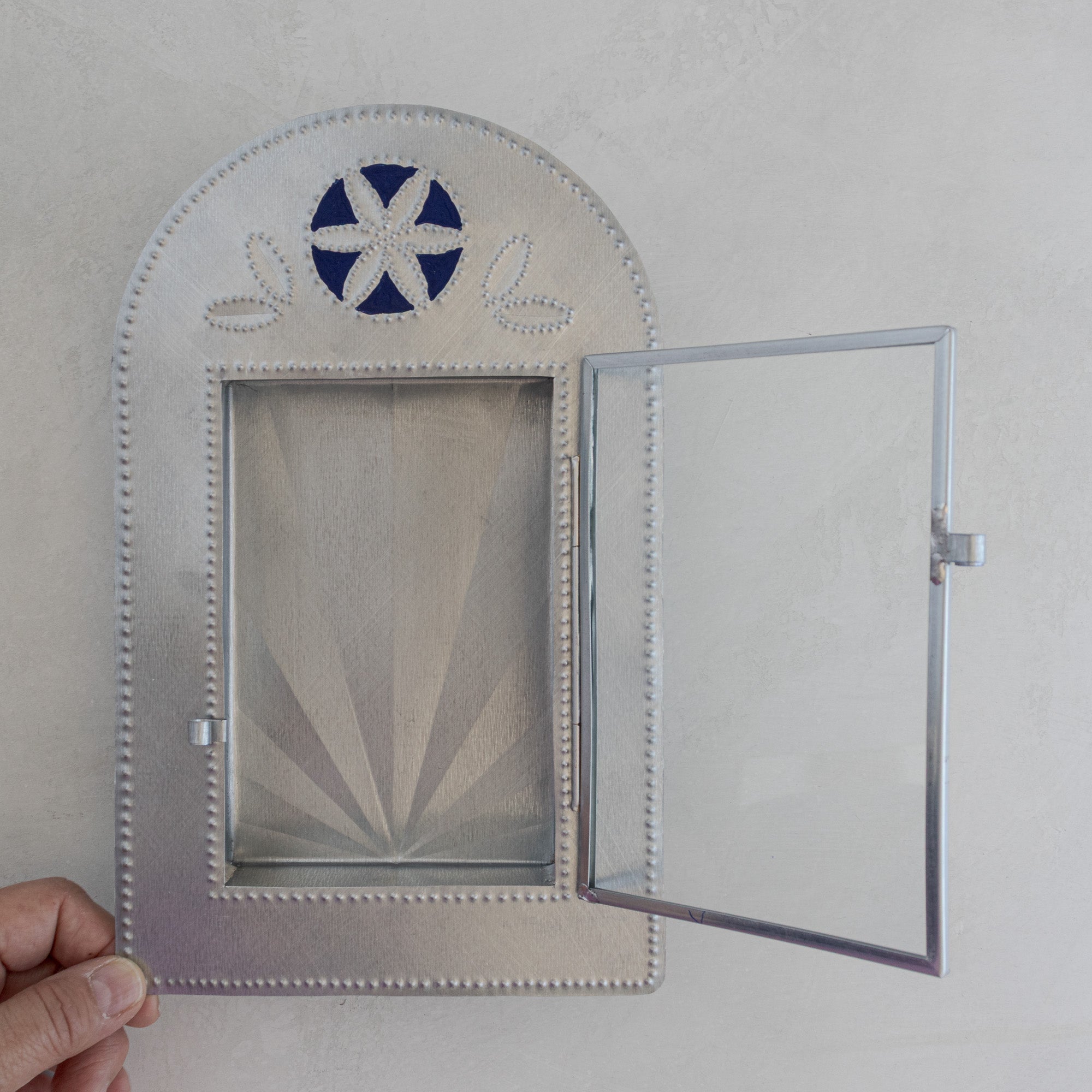 Nicho de Recuerdo held at an angle with glass door fully open, showing interior depth, hinged glass panel, and Prussian blue painted crest motif.