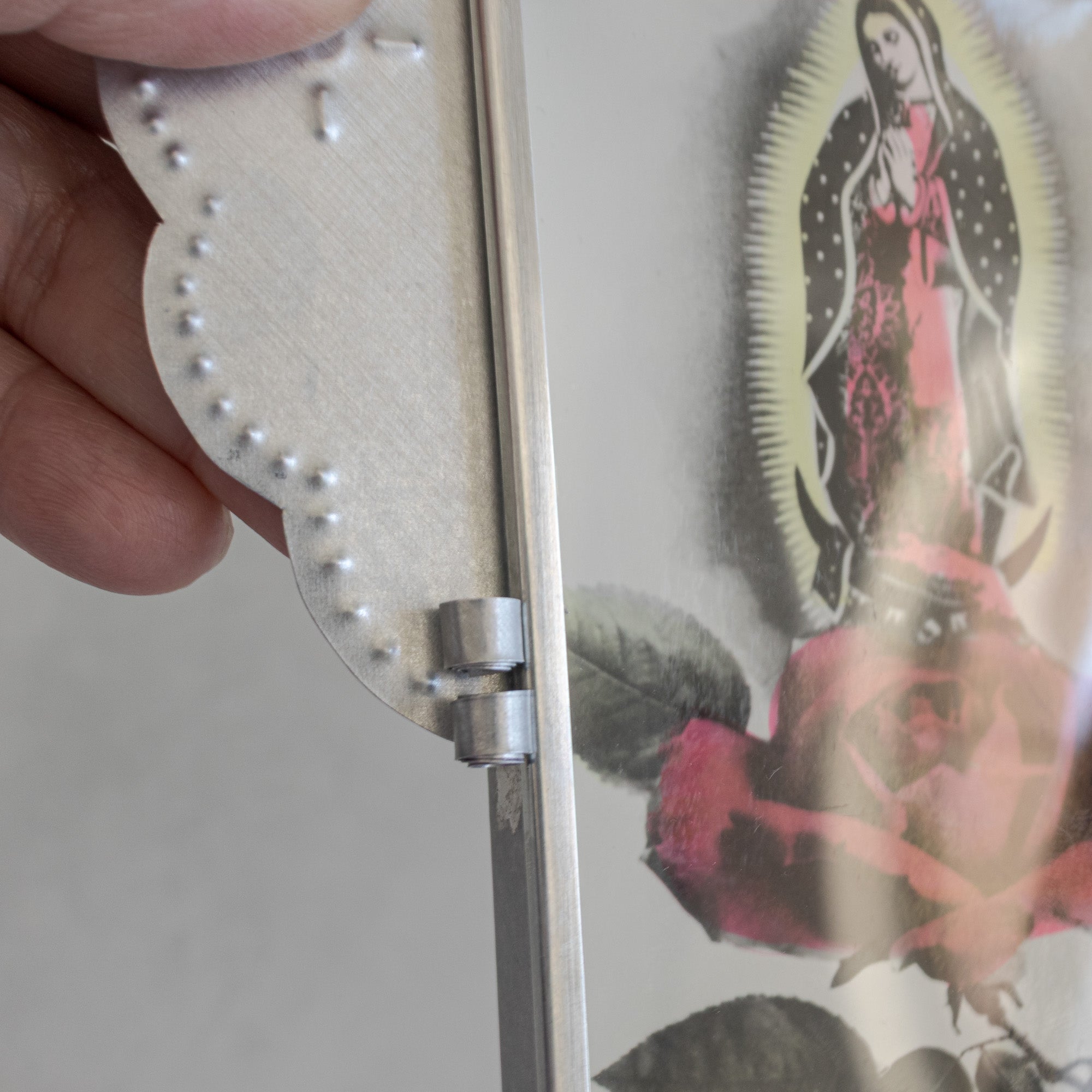 Close-up of Nicho Postal hinge with glass door open, vintage Mexican postcard of La Virgen del Tepeyac visible inside, natural tin body held by hand.