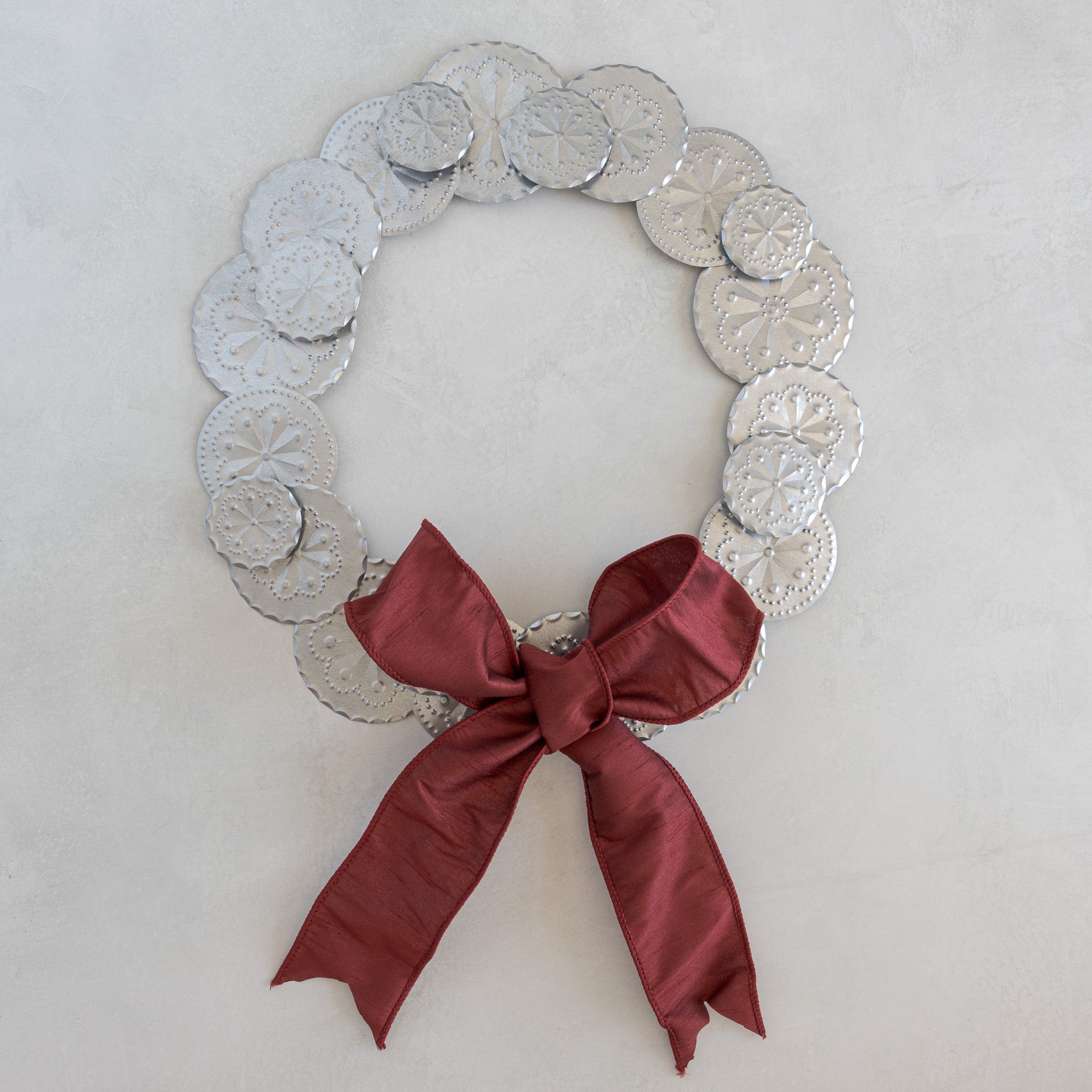 Roseta Corona tinwork wreath with Bordeaux dupioni ribbon bow, hand-punched rosette discs, natural tin, photographed flat on a light plaster surface.