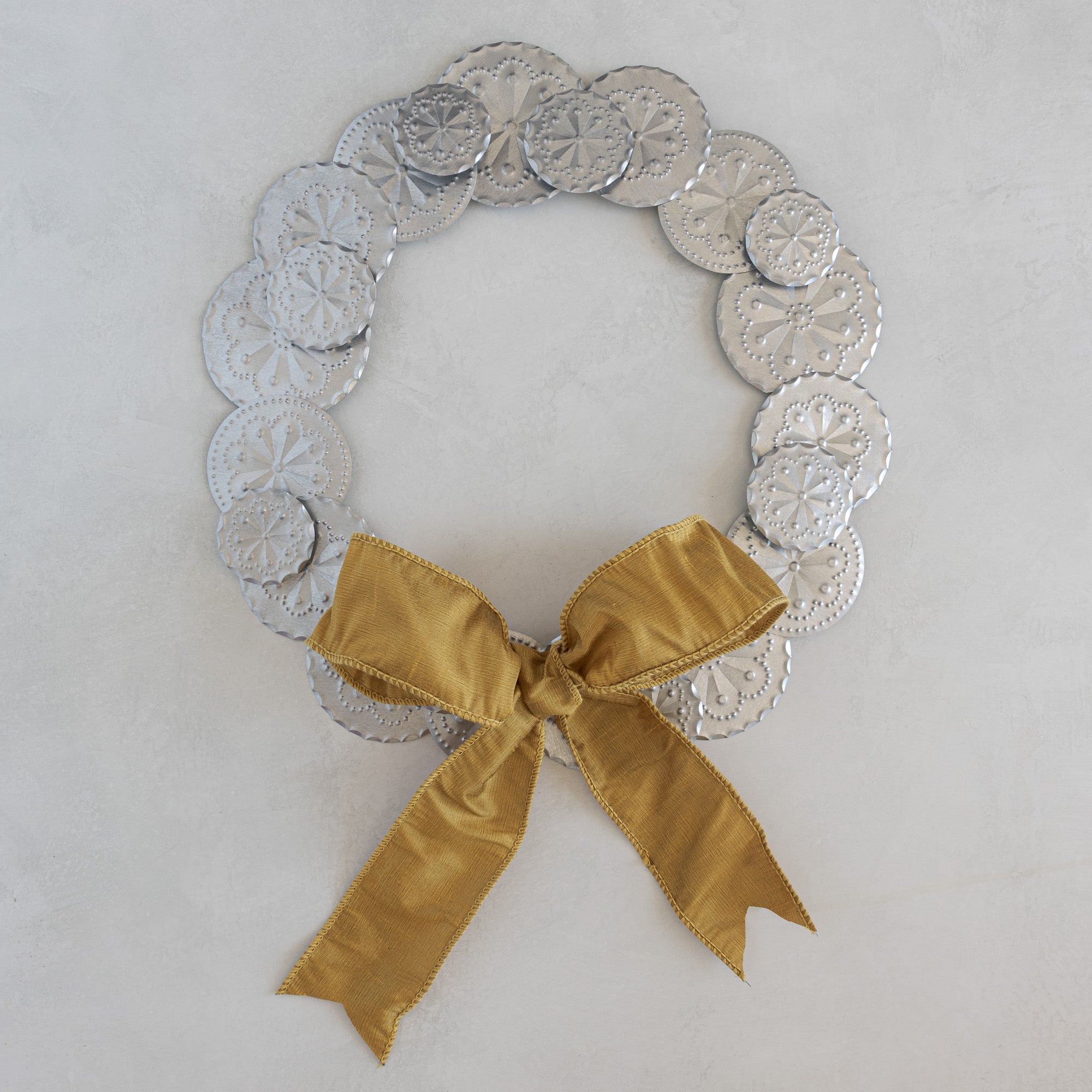 Roseta Corona tinwork wreath with Gold dupioni ribbon bow, hand-punched rosette discs, natural tin, photographed flat on a light plaster surface.