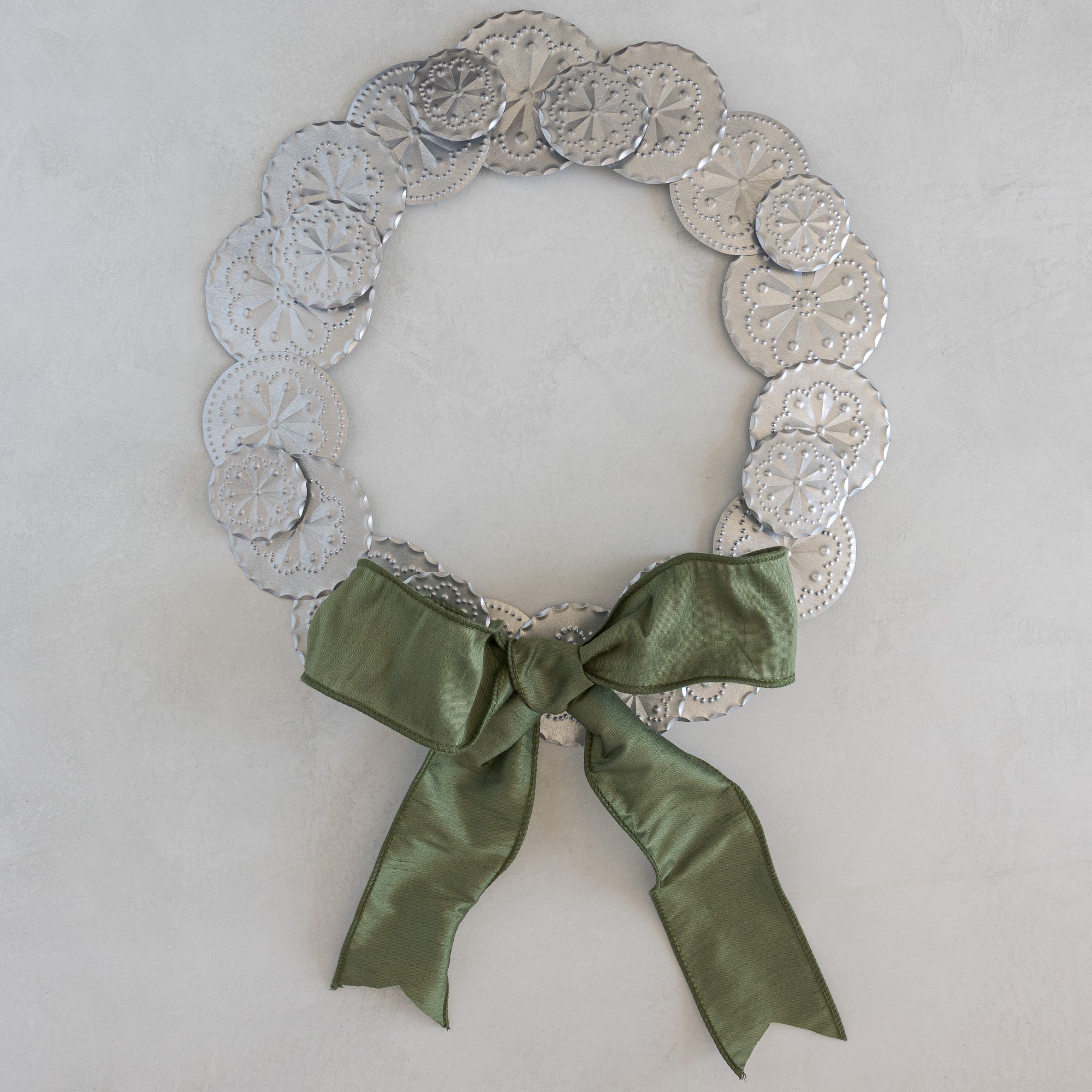 Roseta Corona tinwork wreath with Moss dupioni ribbon bow, hand-punched rosette discs, natural tin, photographed flat on a light plaster surface.