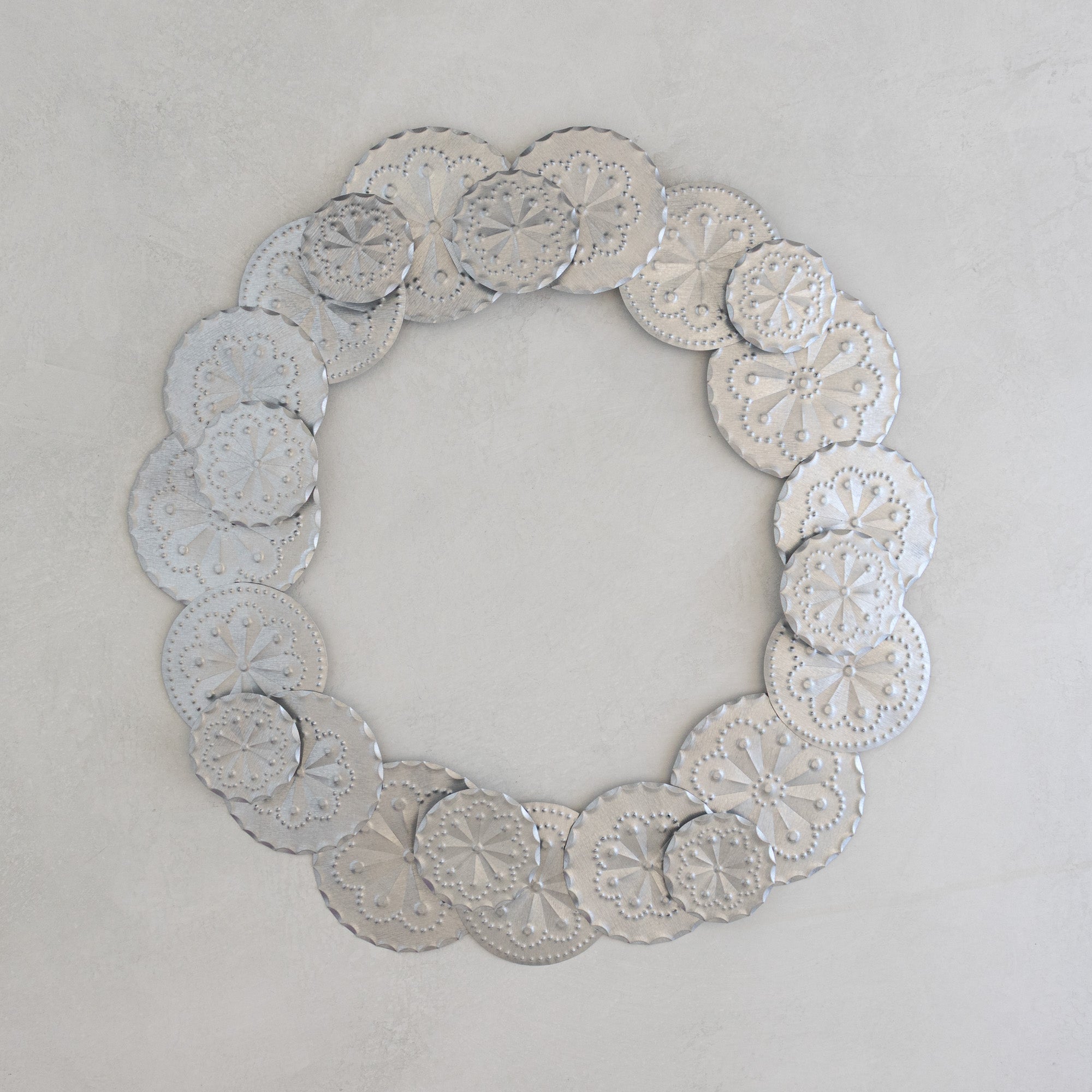 Roseta Corona tinwork wreath in natural tin, hand-punched rosette discs assembled in a circle, photographed flat on a light plaster surface.