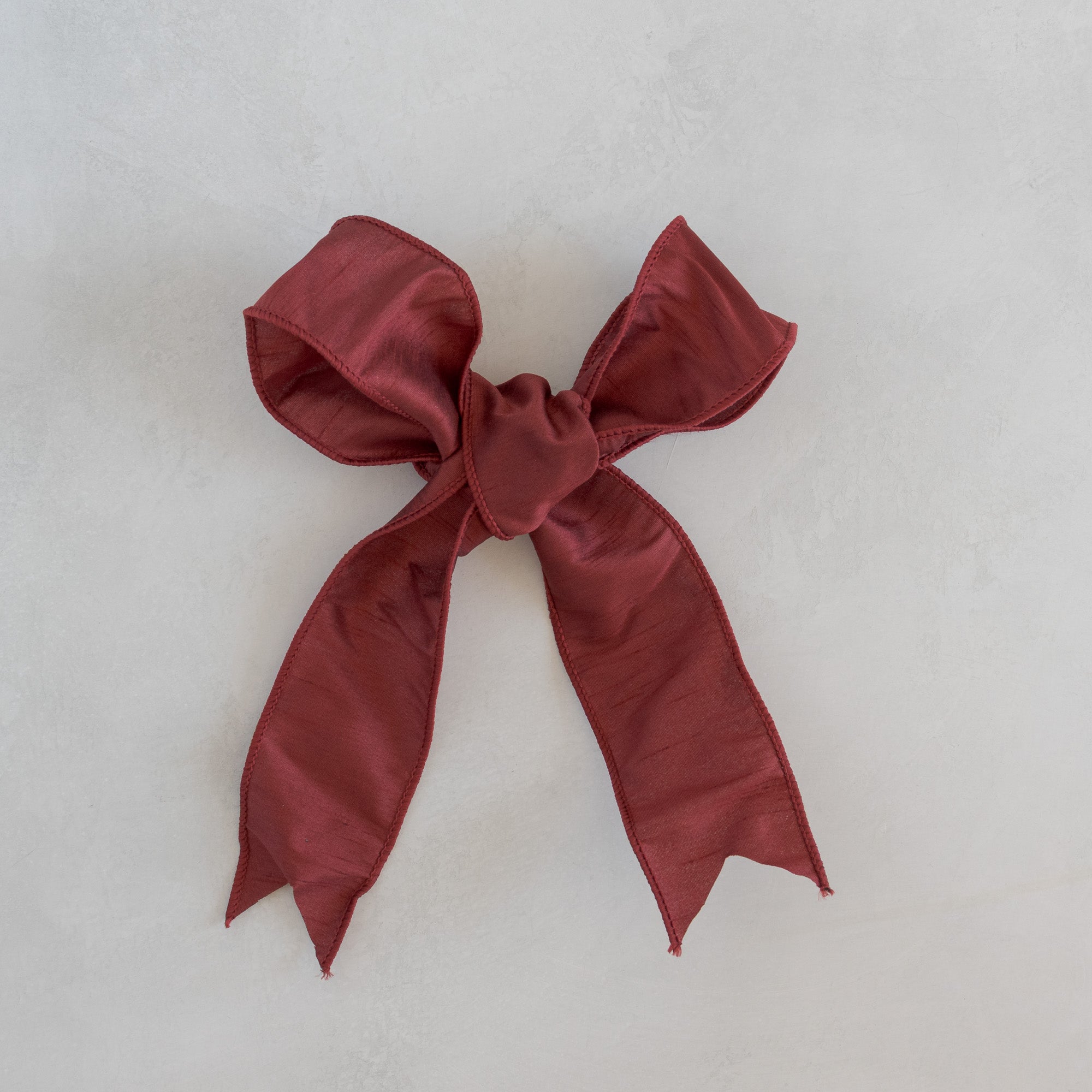 Bordeaux dupioni ribbon bow for Roseta Corona, photographed flat on a light plaster surface.