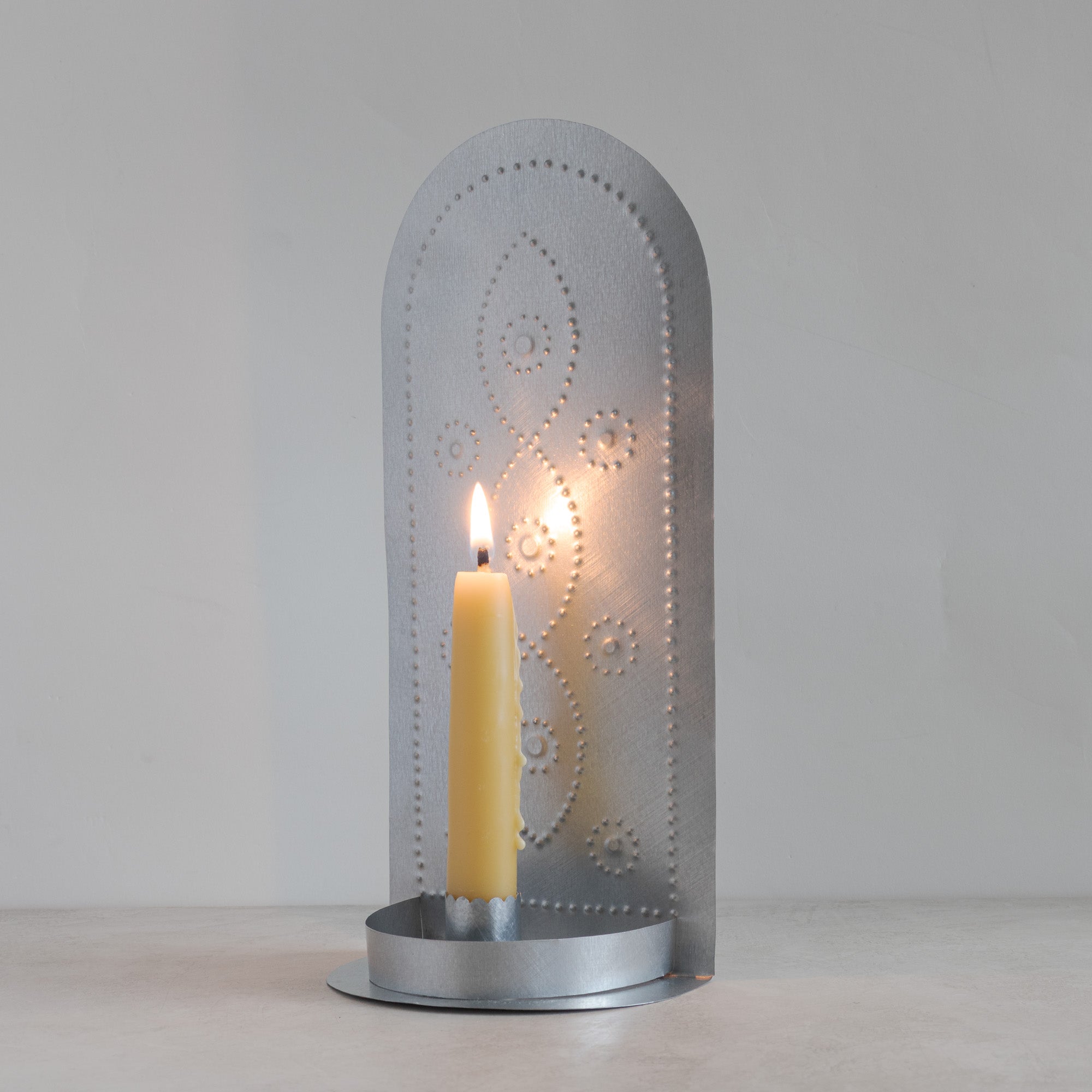Hand-punched tin taper candleholder with vine motif and lit beeswax taper candle on light background